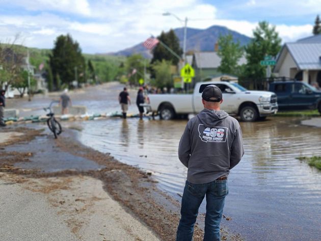 Flooded street with a man in a gray hoodie watching people near a parked truck and bicycles.