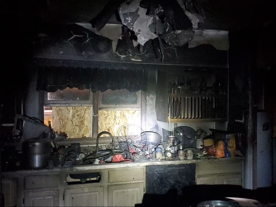 Burned kitchen with charred cabinets, debris, and sunlight through a broken window