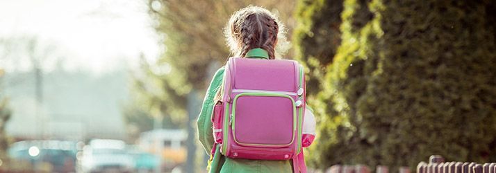 Child with a pink backpack walking outdoors on a sunlit path, viewed from behind.