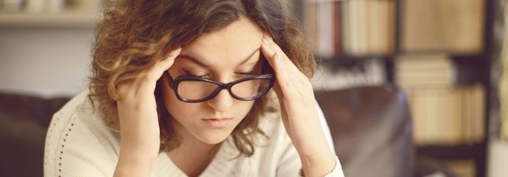 Person in glasses holding their head at a desk, looking stressed or tired, with shelves blurred in the background