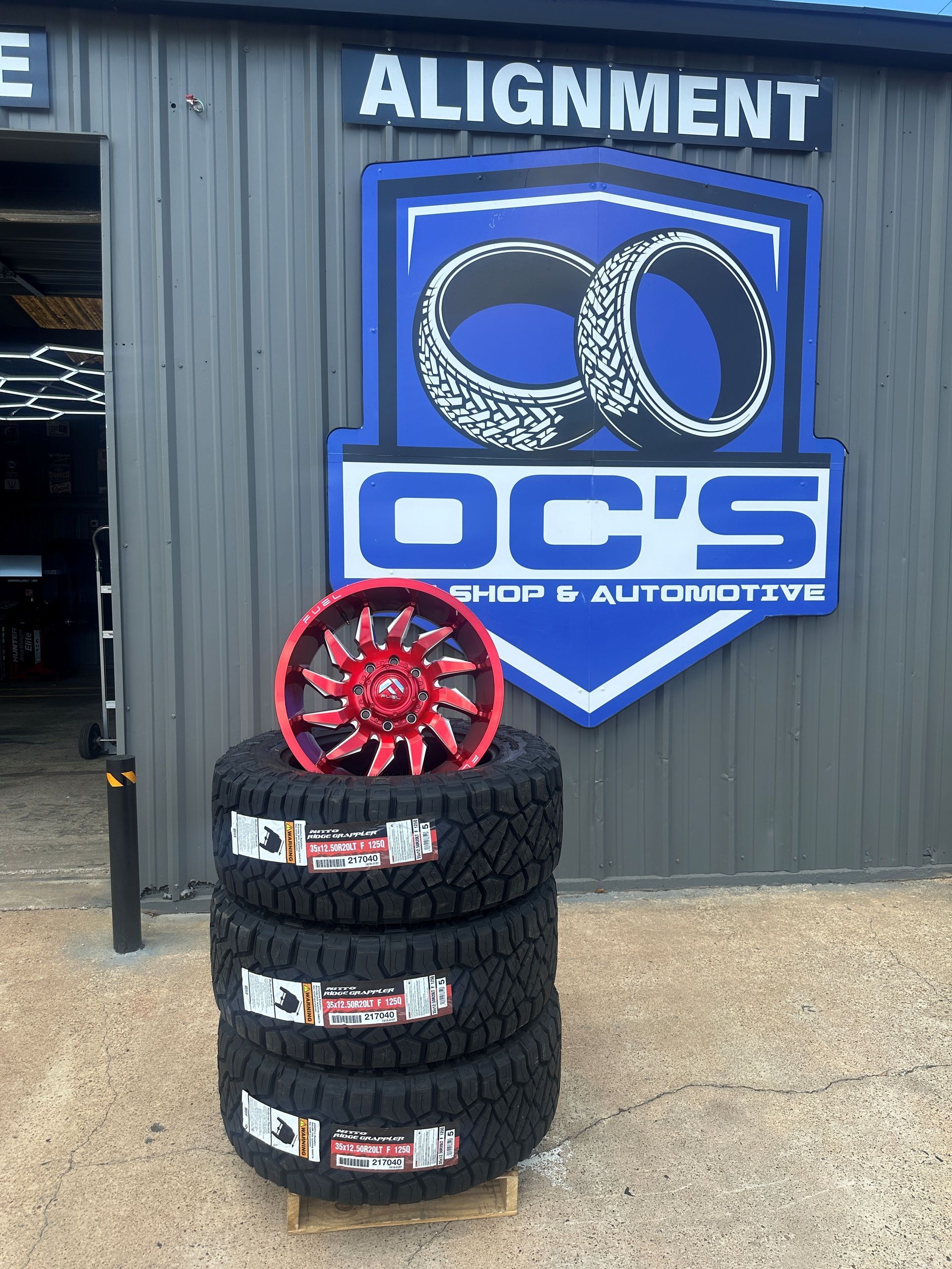 Stacked black tires with a red wheel in front of an OC’s Alignment and Chop & Auto logo sign.