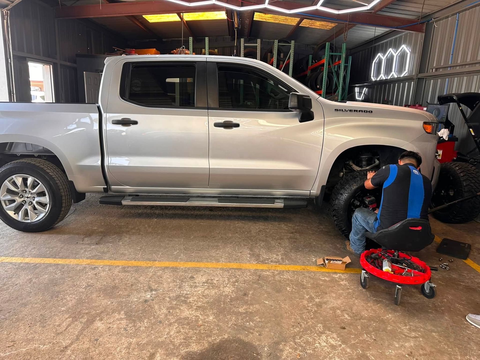Mechanic working on a silver pickup truck in a garage, with tools and a red creeper nearby