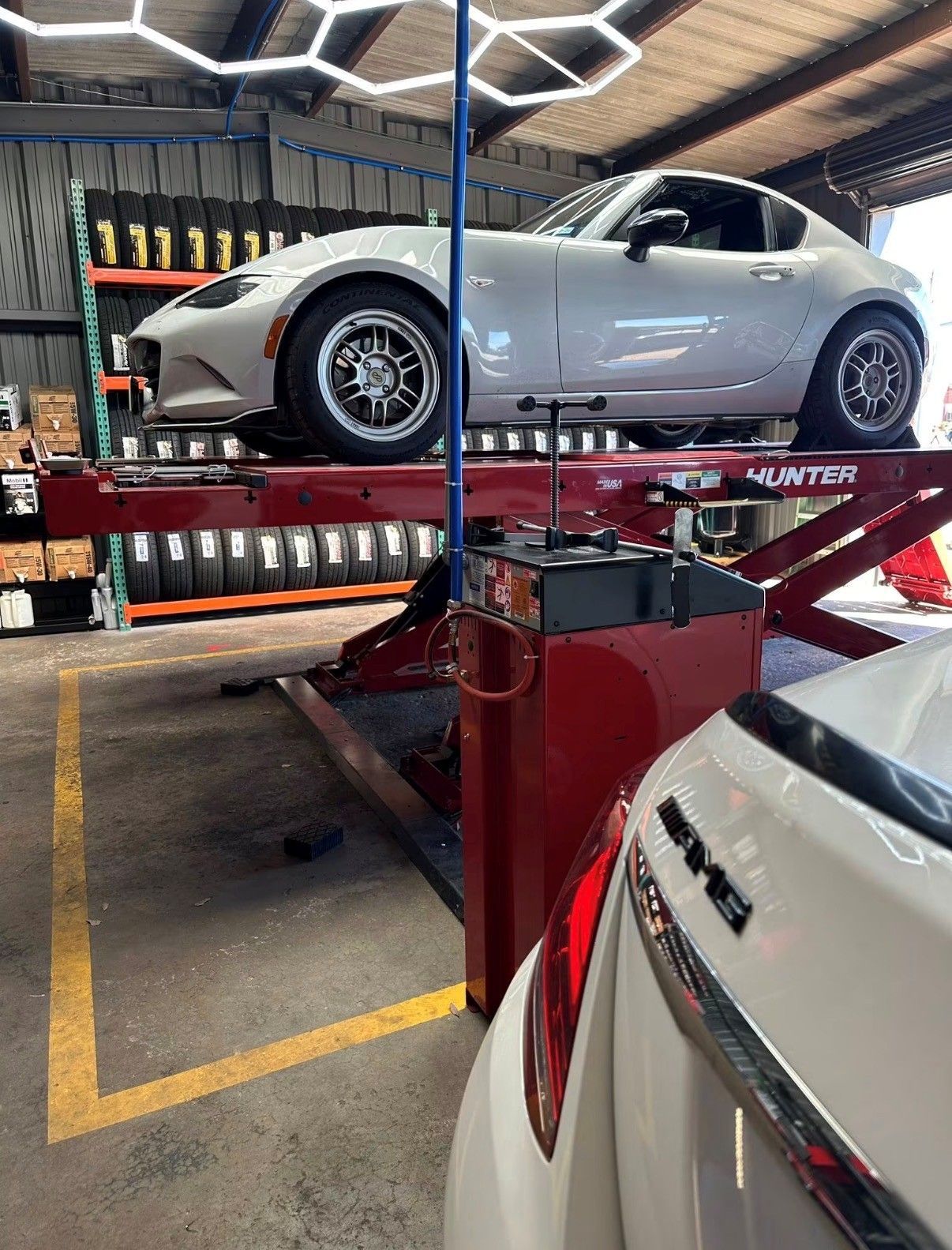 Silver sports car on a red lift in an auto shop, with another white car in the foreground.