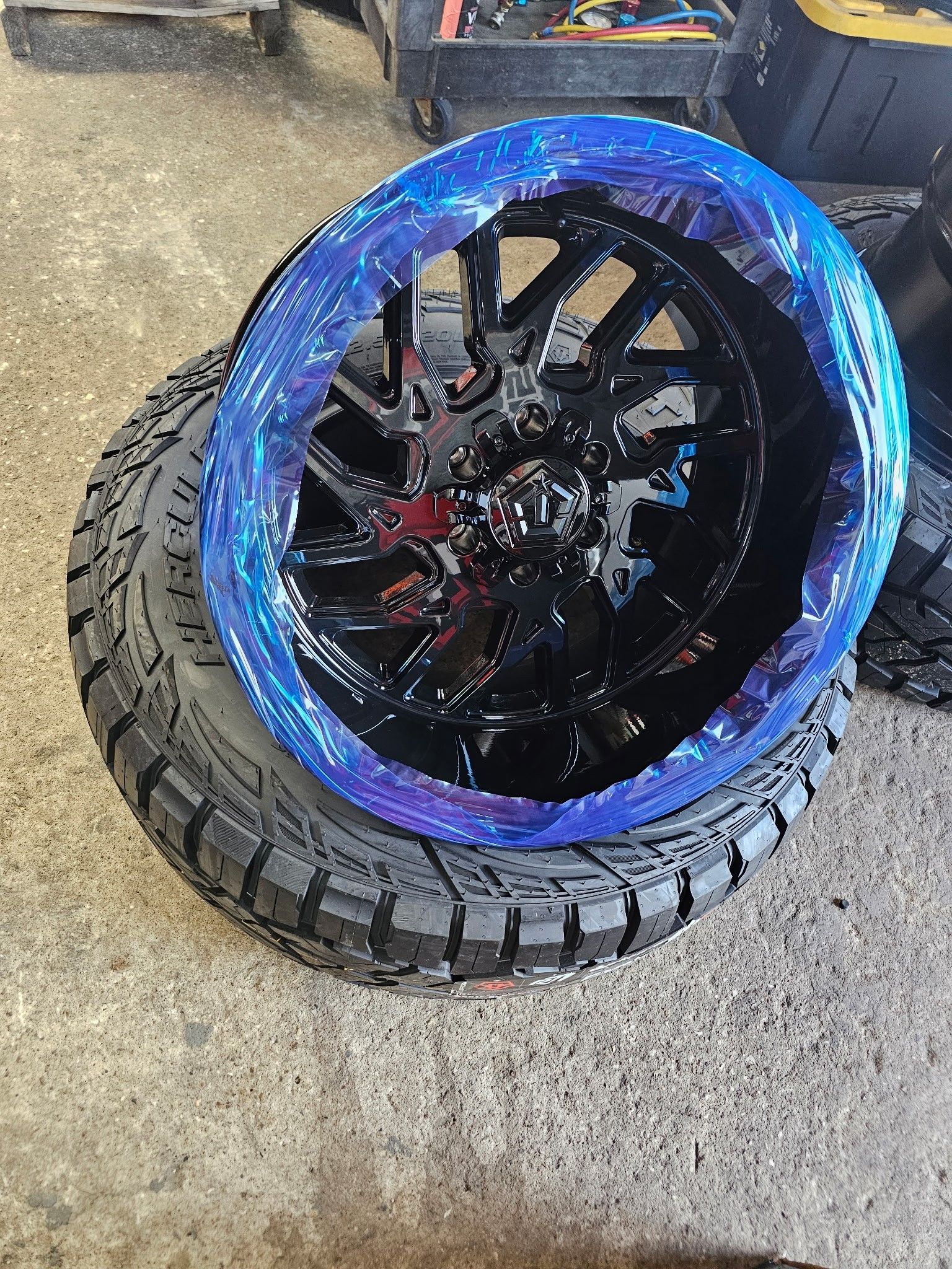 Black off-road tire with a blue rim accent, leaning on another tire on a concrete floor.