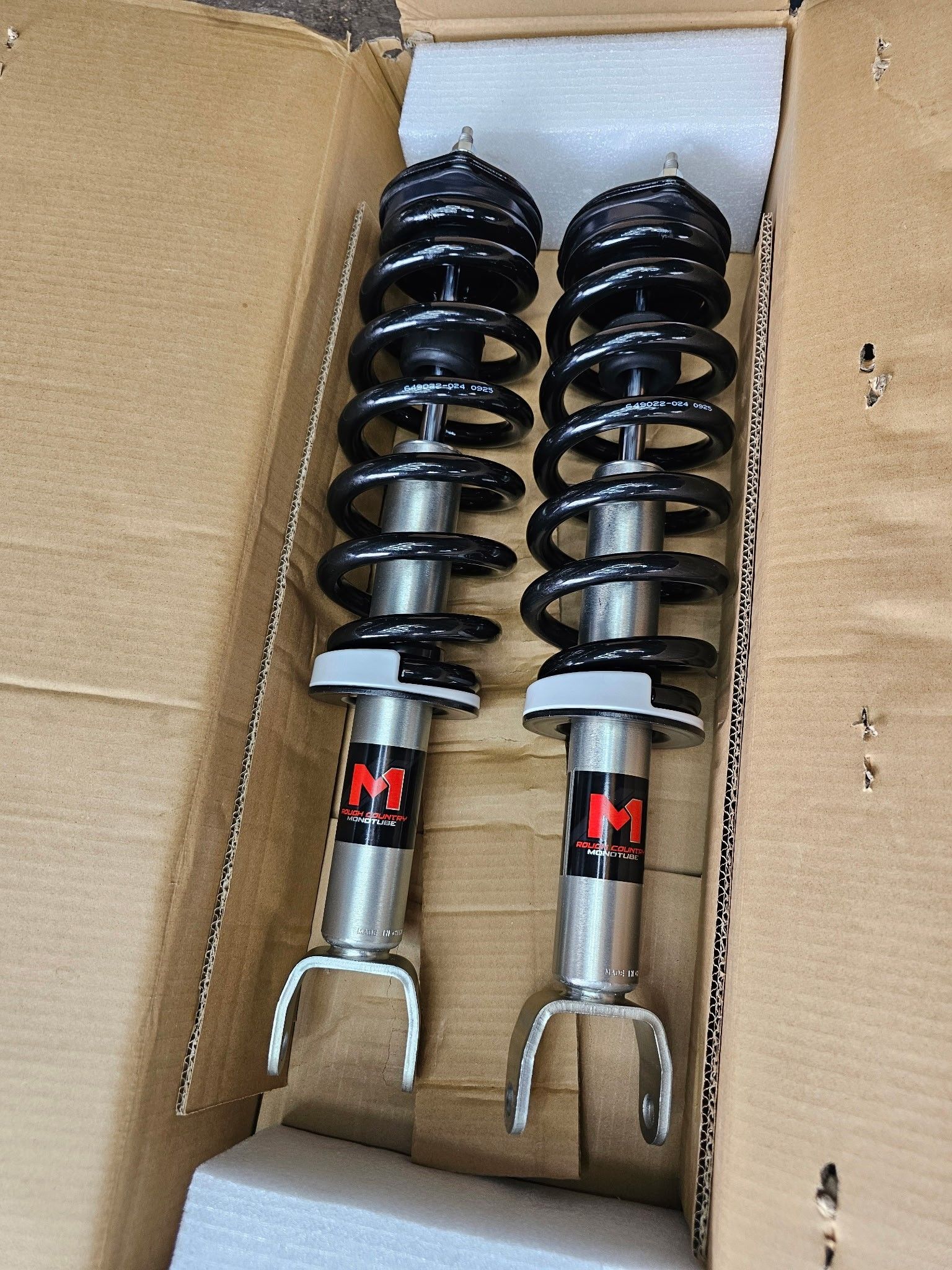 Two black coilover suspension struts in a cardboard box, with silver lower mounts and red labels.