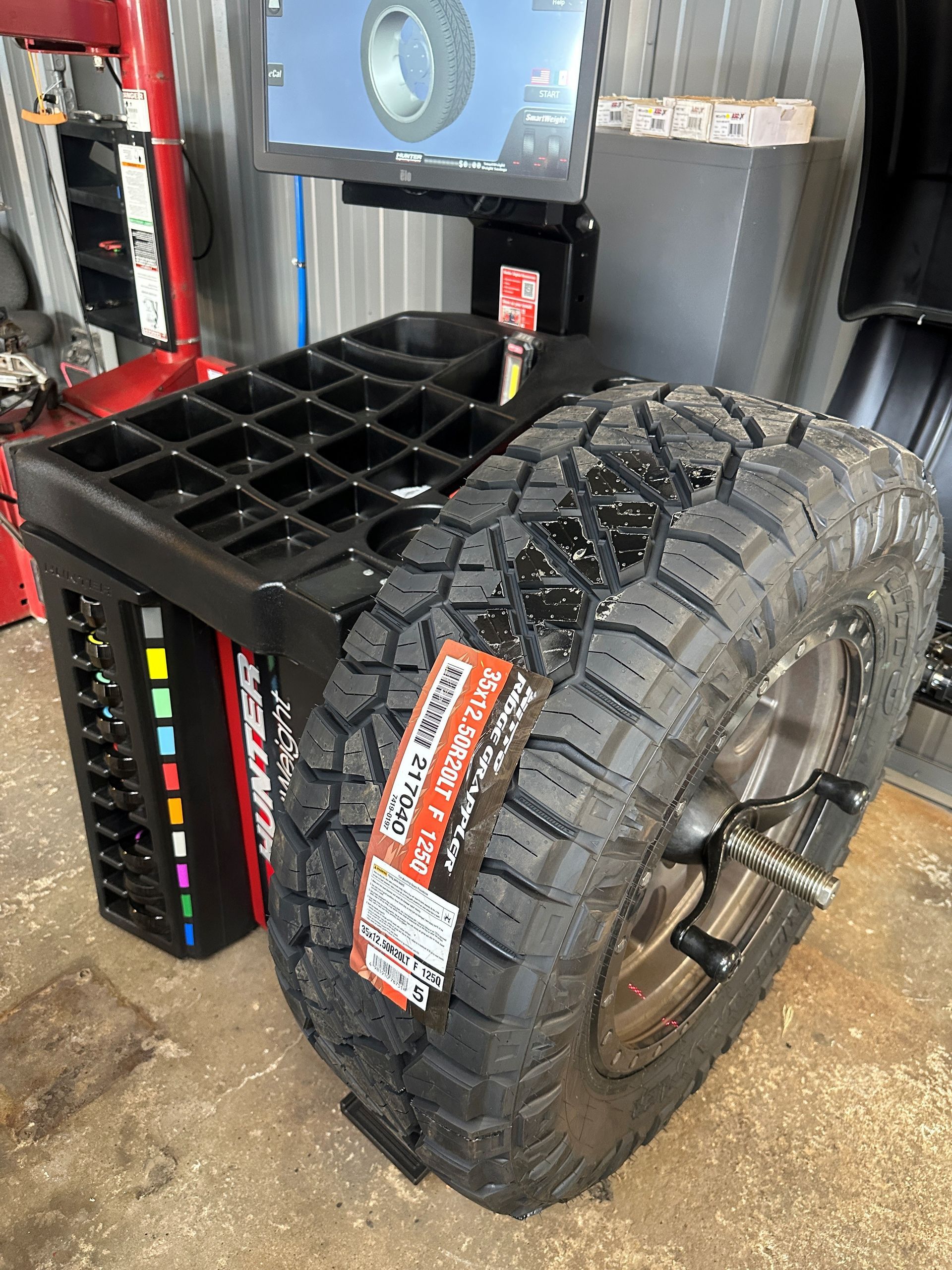 Black off-road tire mounted on a metal rack in an industrial workshop.