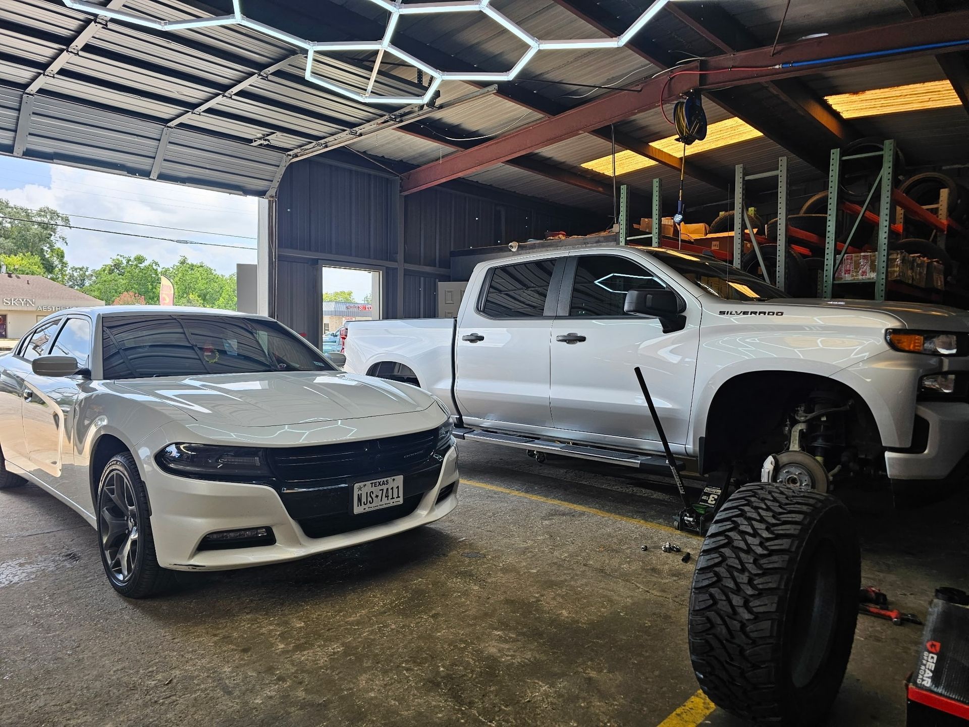 White sedan beside lifted white pickup in a covered garage with large off-road tires in the foreground