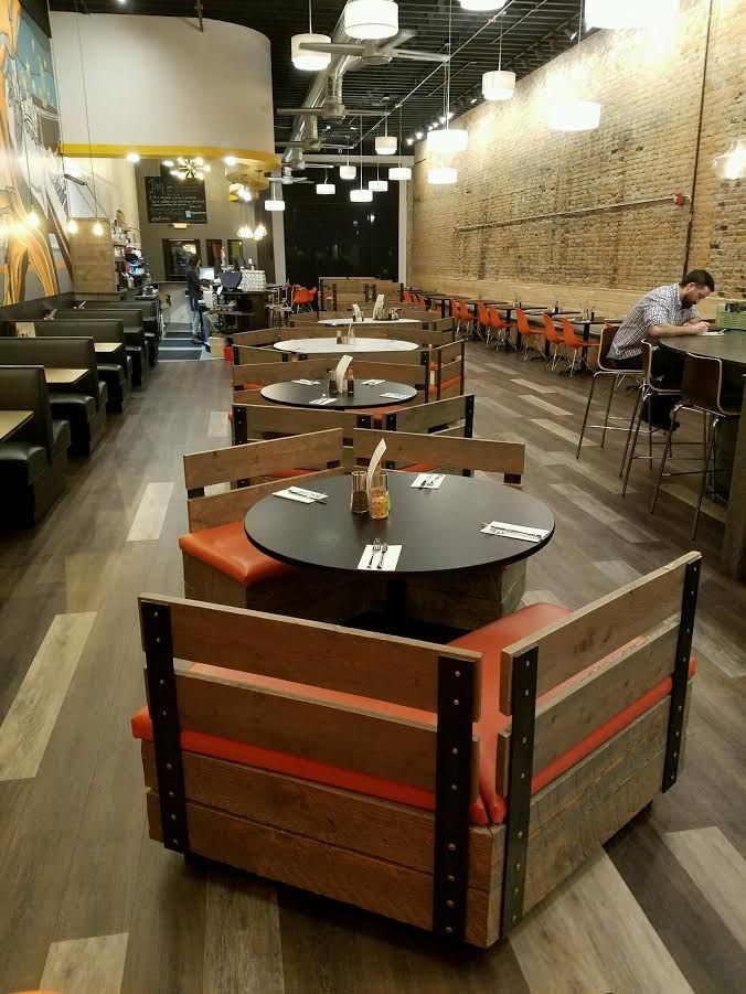 Restaurant interior with wooden booths, dark round tables, and pendant lighting along a textured brick wall.