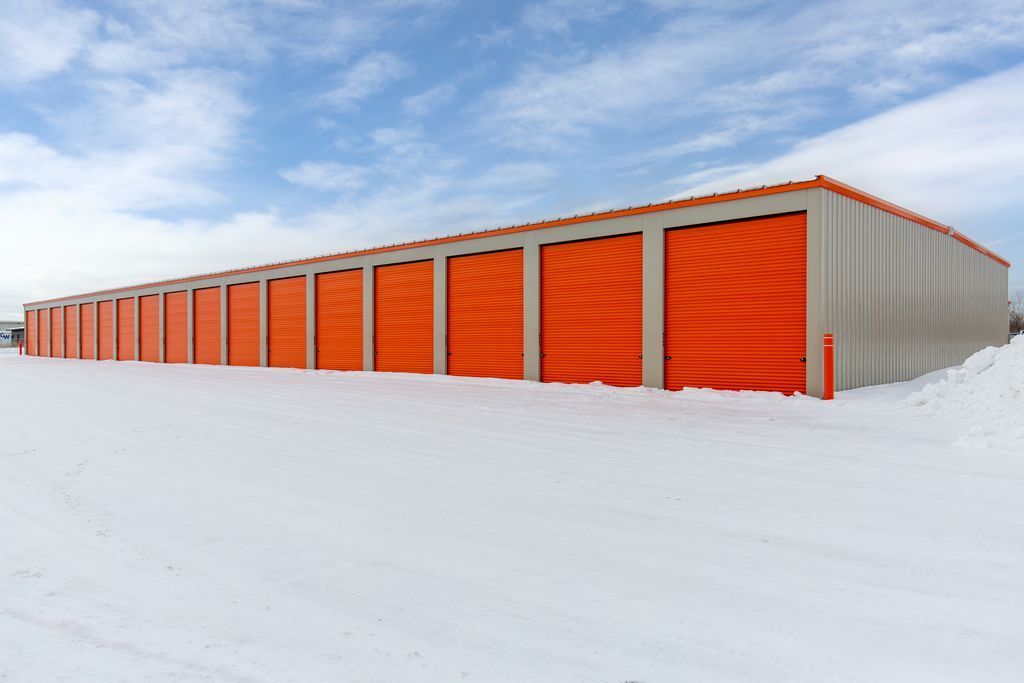 A row of storage units with bright orange roll-up doors, set against a snow-covered ground and a blue sky with clouds.