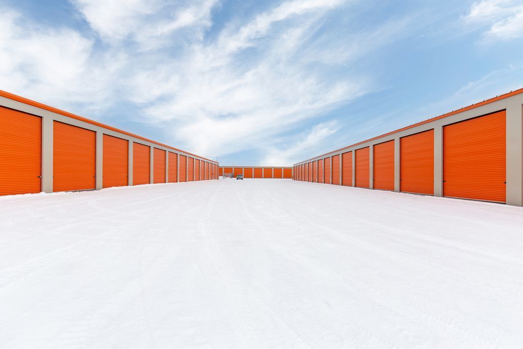 Rows of orange roll-up storage unit doors line both sides of a wide, empty paved lot under a blue sky with light clouds.