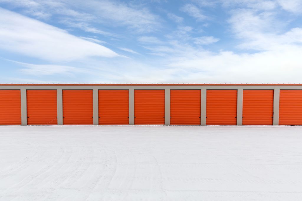 A row of bright orange self-storage unit doors set against a white, snow-covered ground under a blue sky.