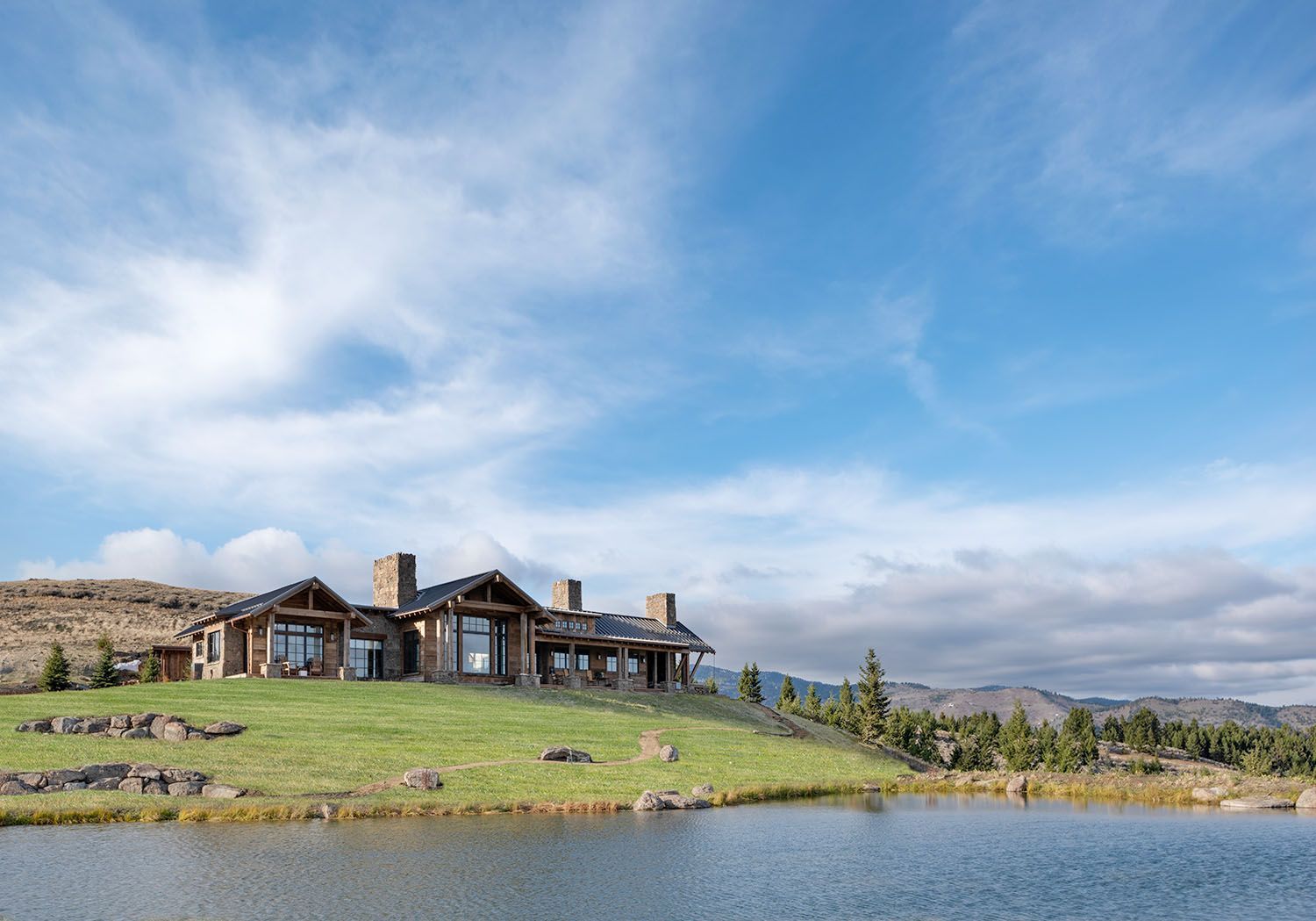 A rustic stone and wood house sits on a grassy hill above a calm pond under a cloudy blue sky