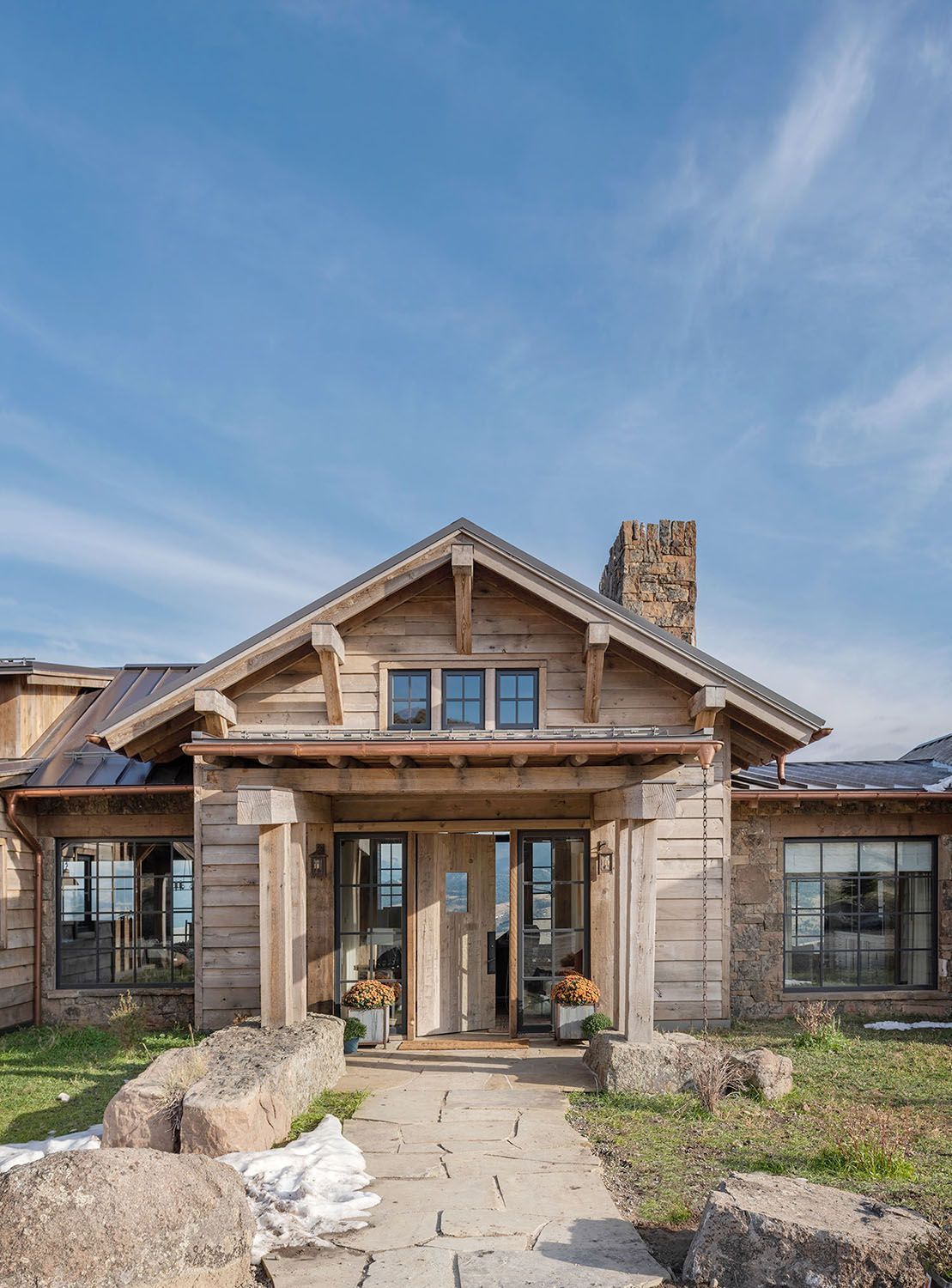 A rustic wood-and-stone cabin entrance featuring a covered porch, stone columns, and large windows under a blue sky