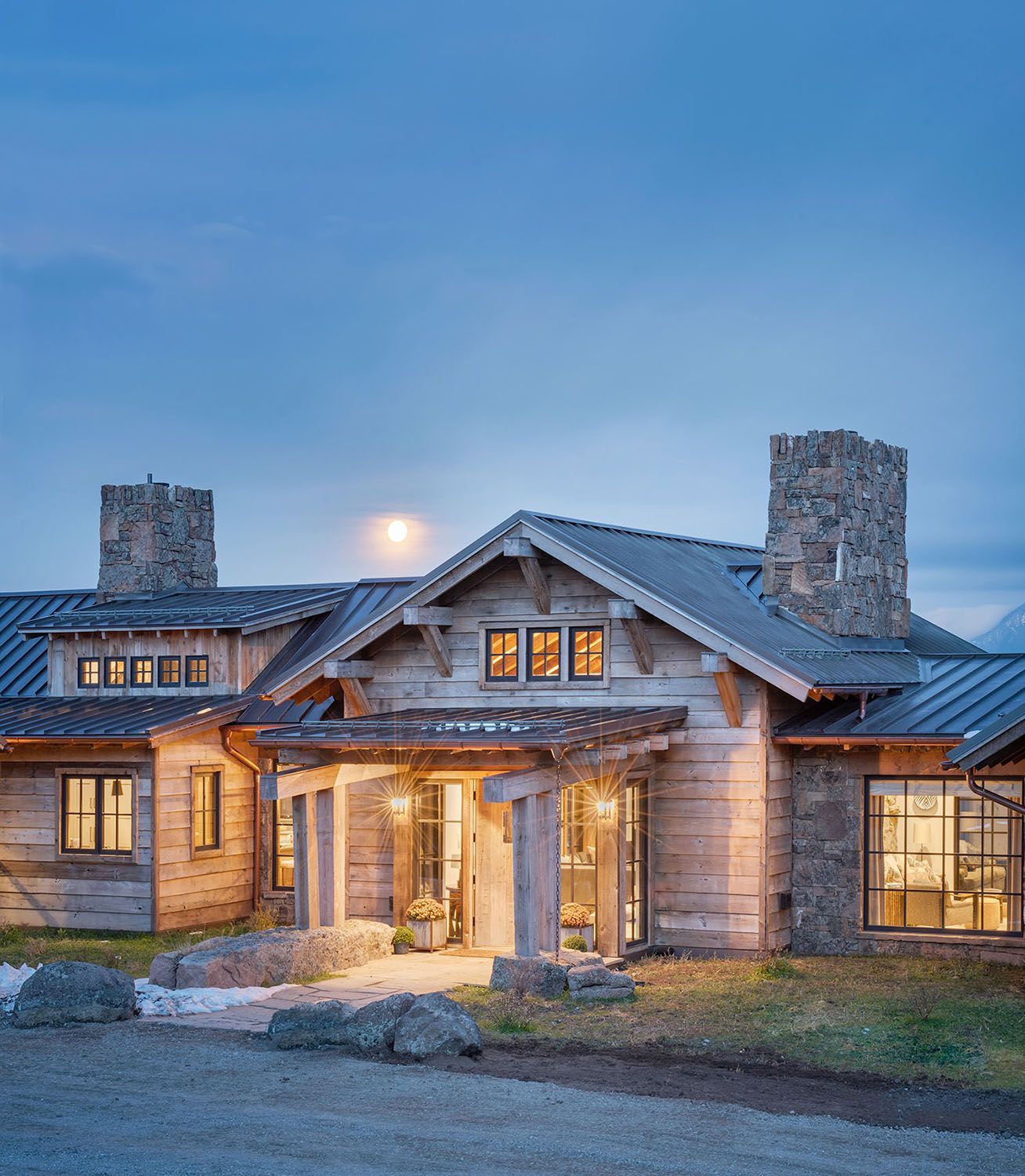 A rustic wood and stone house with a metal roof and glowing windows at dusk