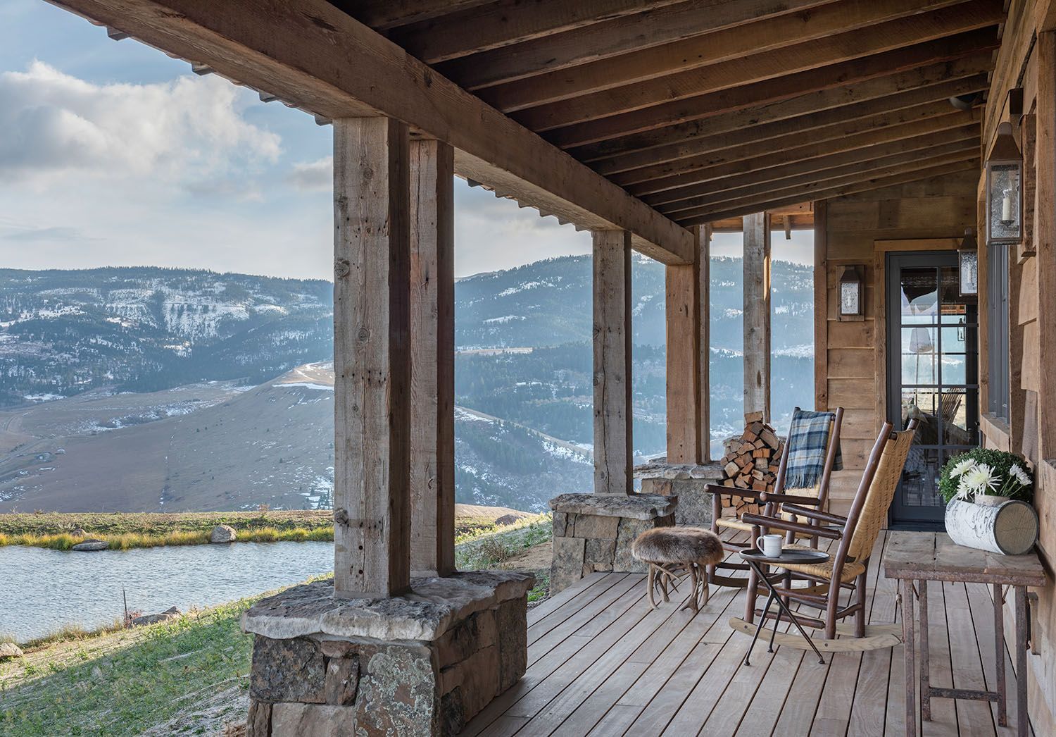 A wooden porch with rustic rocking chairs overlooking a scenic valley with snow-capped mountains and a pond