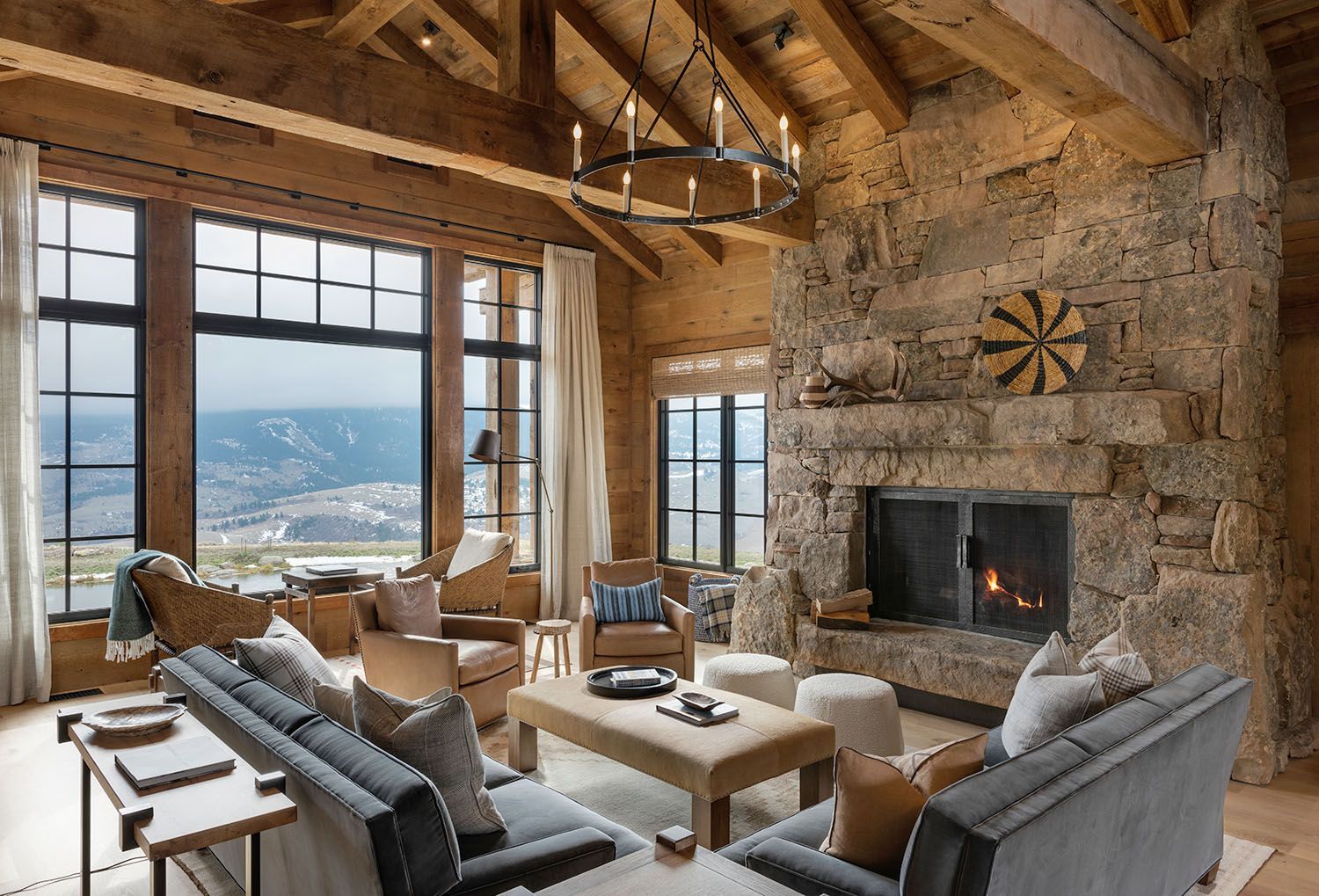 A cozy living room with wooden vaulted ceilings, a large stone fireplace, and a wall of windows overlooking a mountain