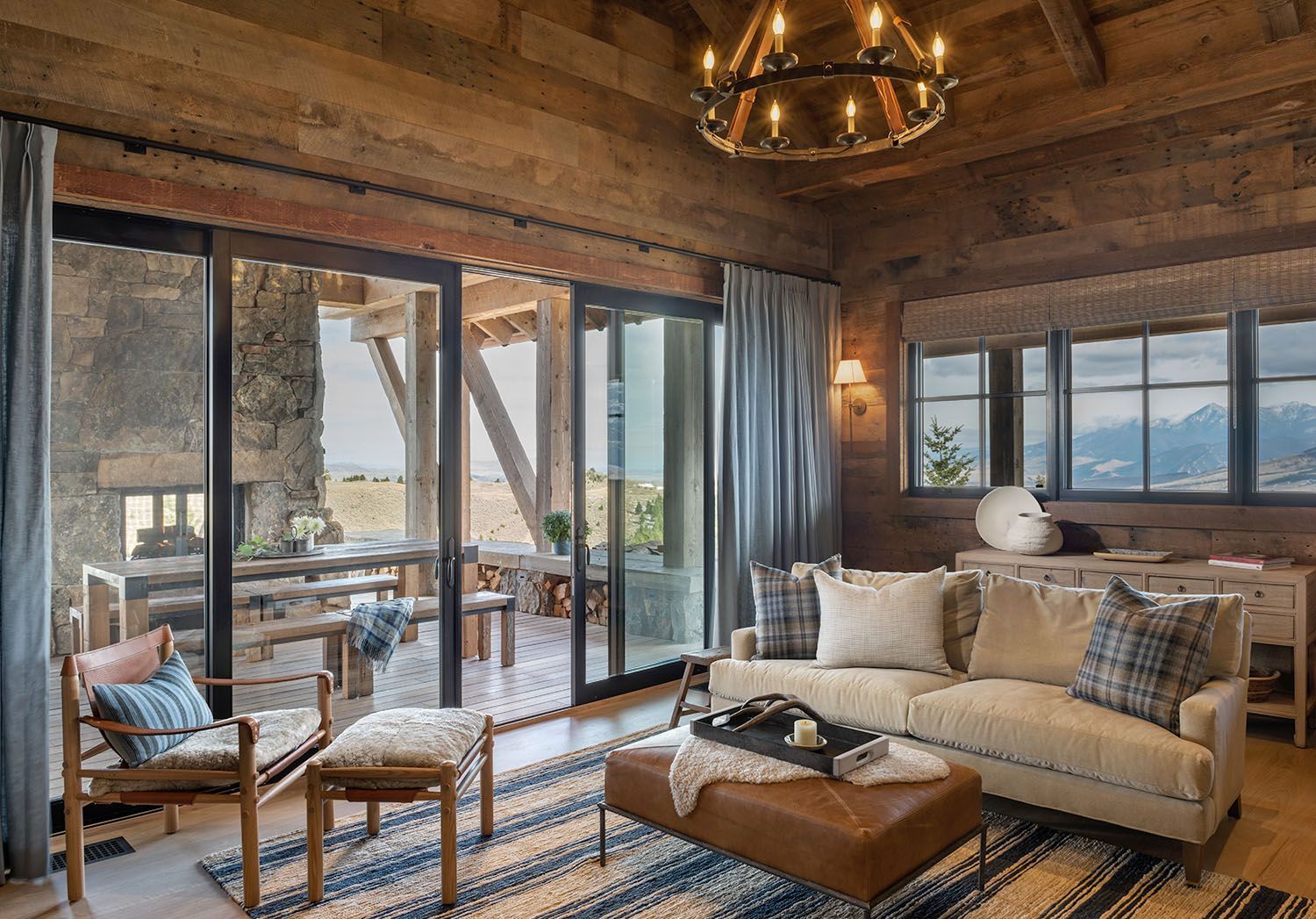 A rustic living room with wood-paneled walls, a cream sofa, leather ottoman, and large glass doors opening to a patio