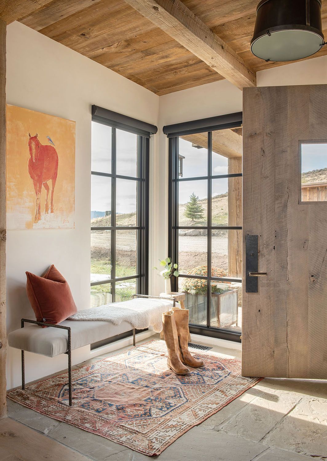 A cozy entryway with a rustic wooden door, a bench, a patterned rug, a horse painting, and large black-framed windows