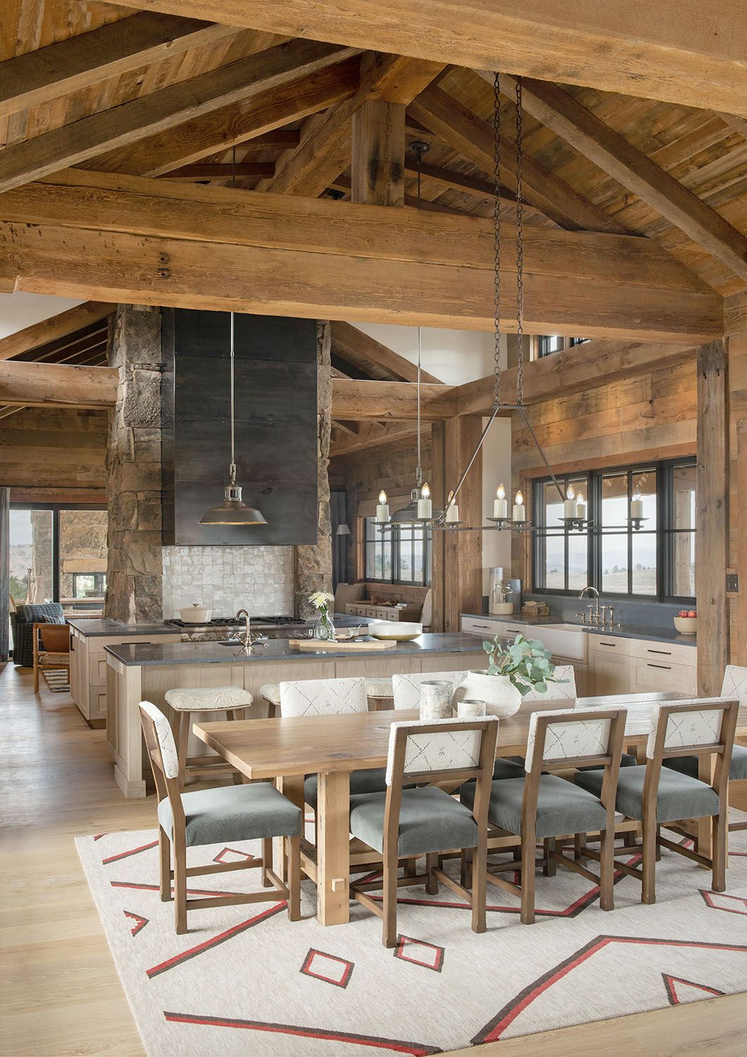 A rustic dining area with wooden beams, a long table with grey chairs, and an open kitchen with a black hood