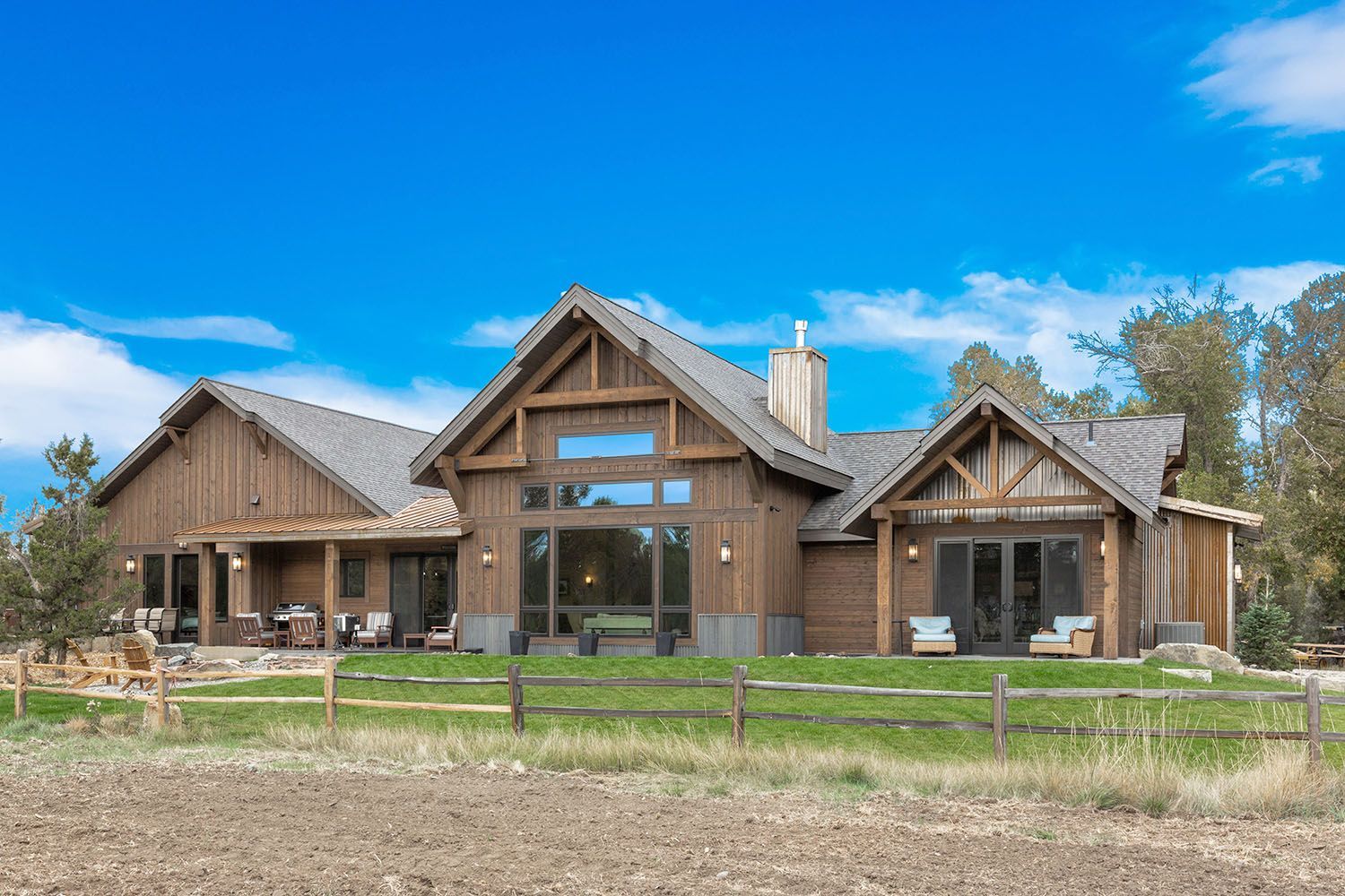 A wood-sided home with a gray roof and large windows sits behind a wooden fence under a bright blue sky