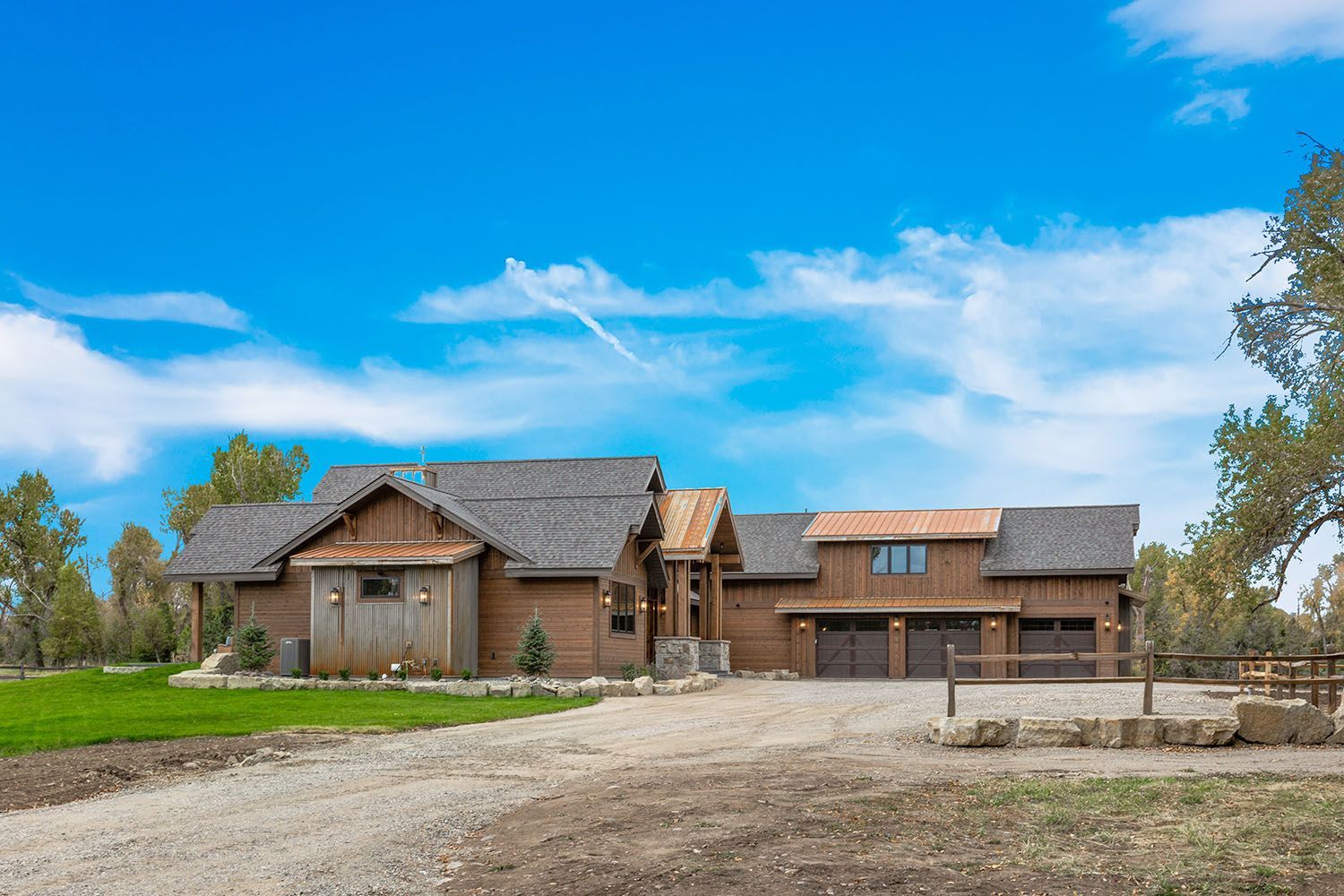 A rustic wood-sided house with a gravel driveway, gray shingled roof, and three-car garage under a bright blue sky