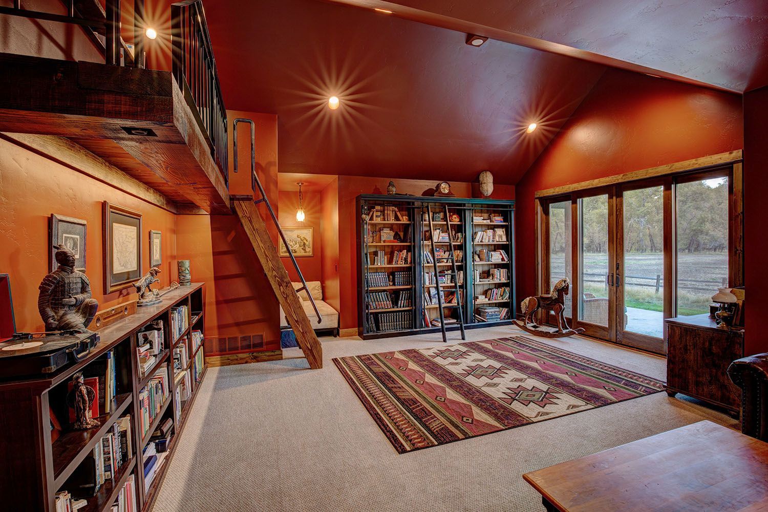 A warm-toned room with vaulted ceilings, large bookshelves, a wooden loft ladder, patterned rug, and glass patio doors
