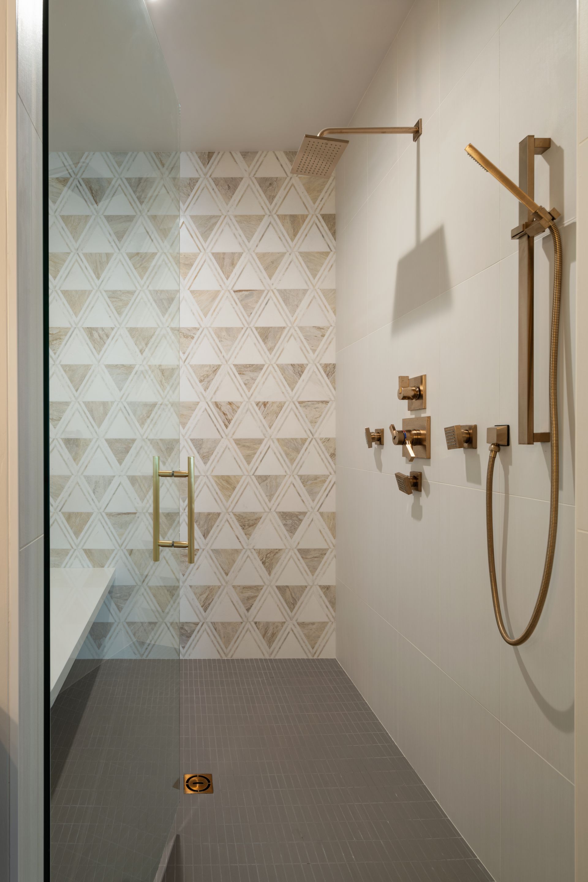 A modern walk-in shower with gold fixtures, a white-tiled wall with a geometric pattern, and a grey tile floor.