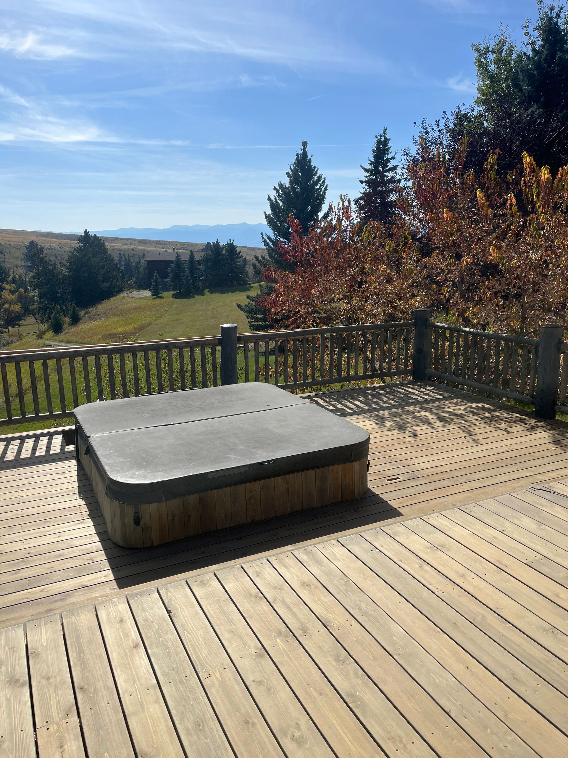 A hot tub with a closed cover sits on a wooden deck overlooking a grassy hill, evergreen trees, and distant mountains.