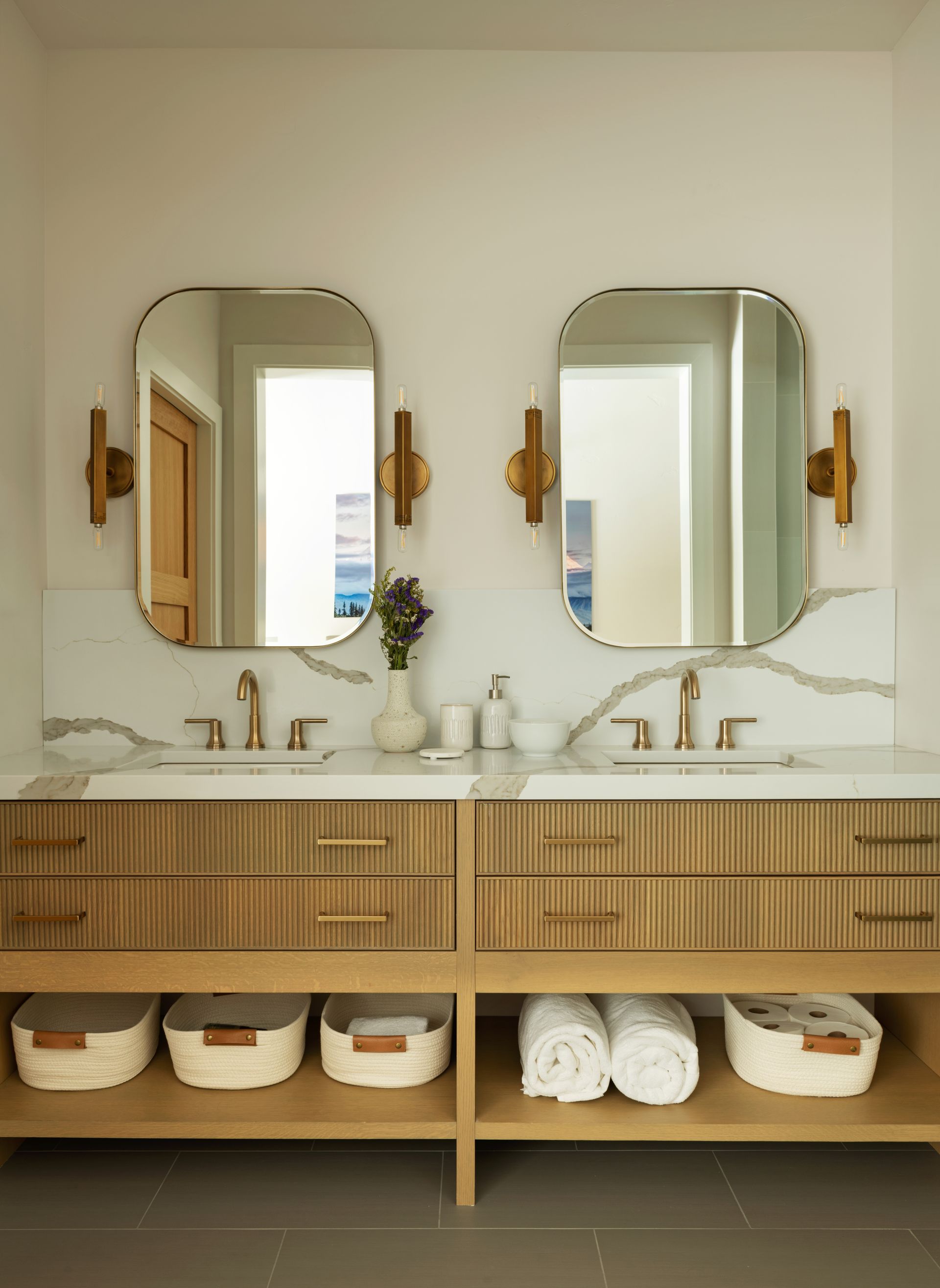 A double bathroom vanity with slatted wood drawers, marble backsplash, two mirrors, gold sconces, and woven storage bins.