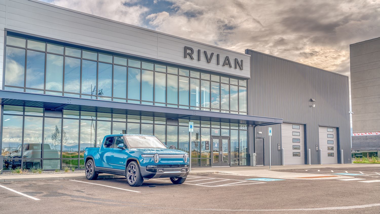 Blue Rivian electric truck parked in front of a Rivian dealership. Building with large windows, overcast sky.