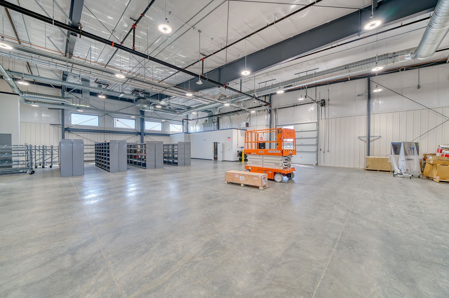 Large, empty warehouse with concrete floor. Orange lift equipment, shelving, and a loading door are visible.