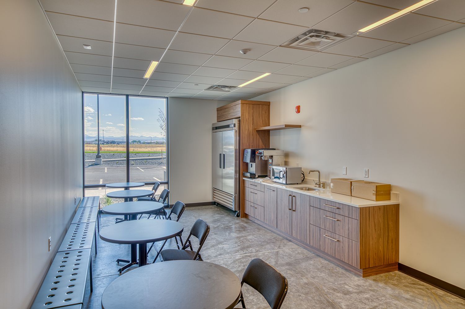 Modern break room with tables, chairs, coffee station, refrigerator, and large window overlooking a landscape.