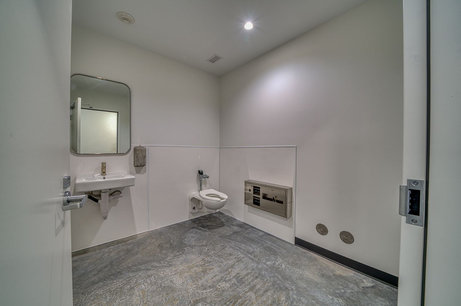 A minimalist, empty public restroom with a toilet, sink, and mirror. White walls, gray floor.