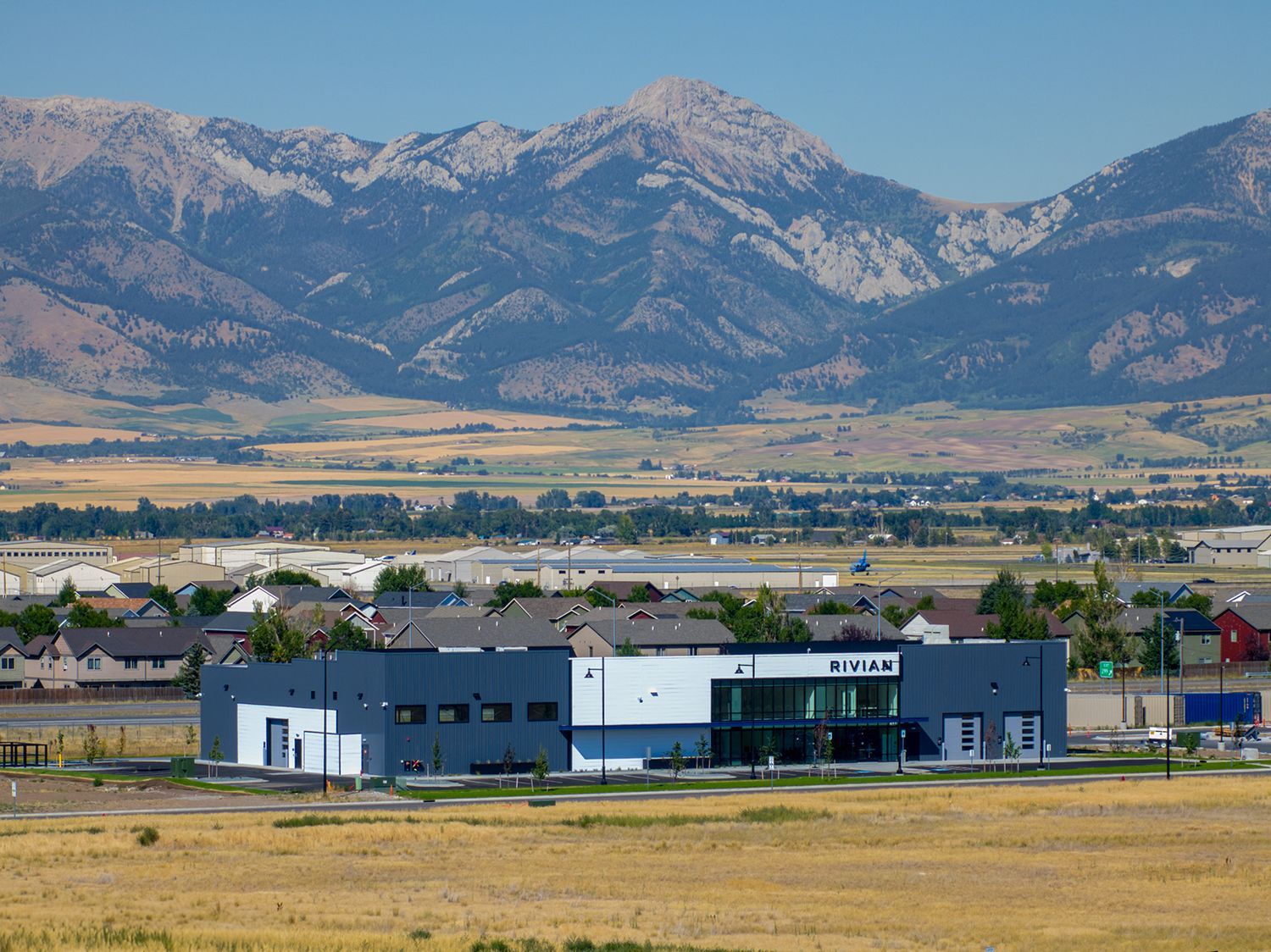 Modern building with glass front and dark grey accents, fields, houses, and mountains in background.