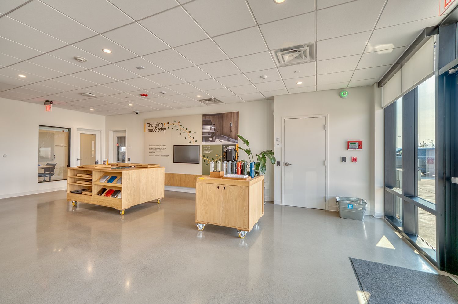 Modern, brightly-lit interior space with display tables and large windows. Concrete floors, white walls, and various products on display.