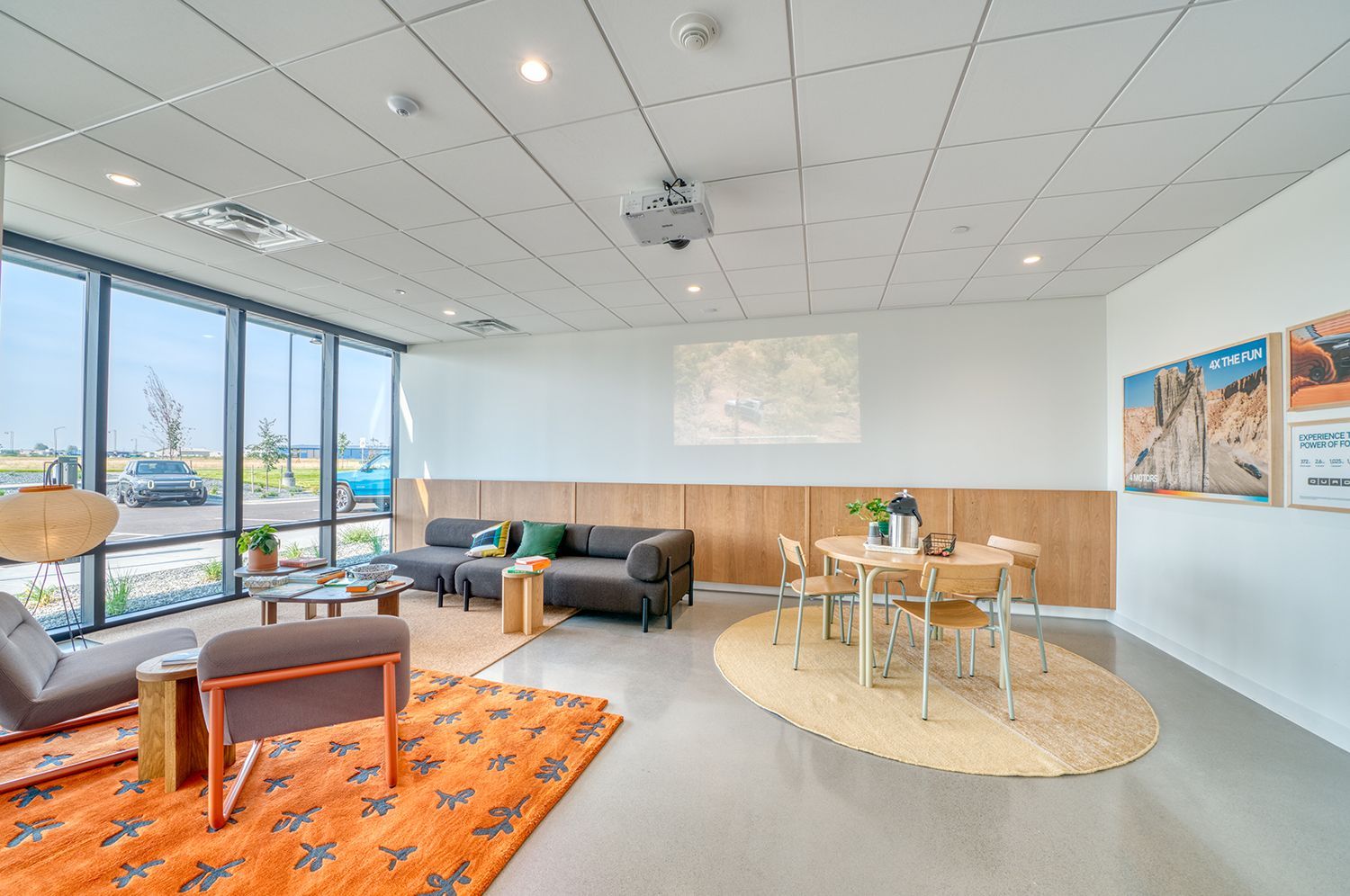 A modern office lounge with seating, a small round table, and a projector screen. Large windows offer an outside view.