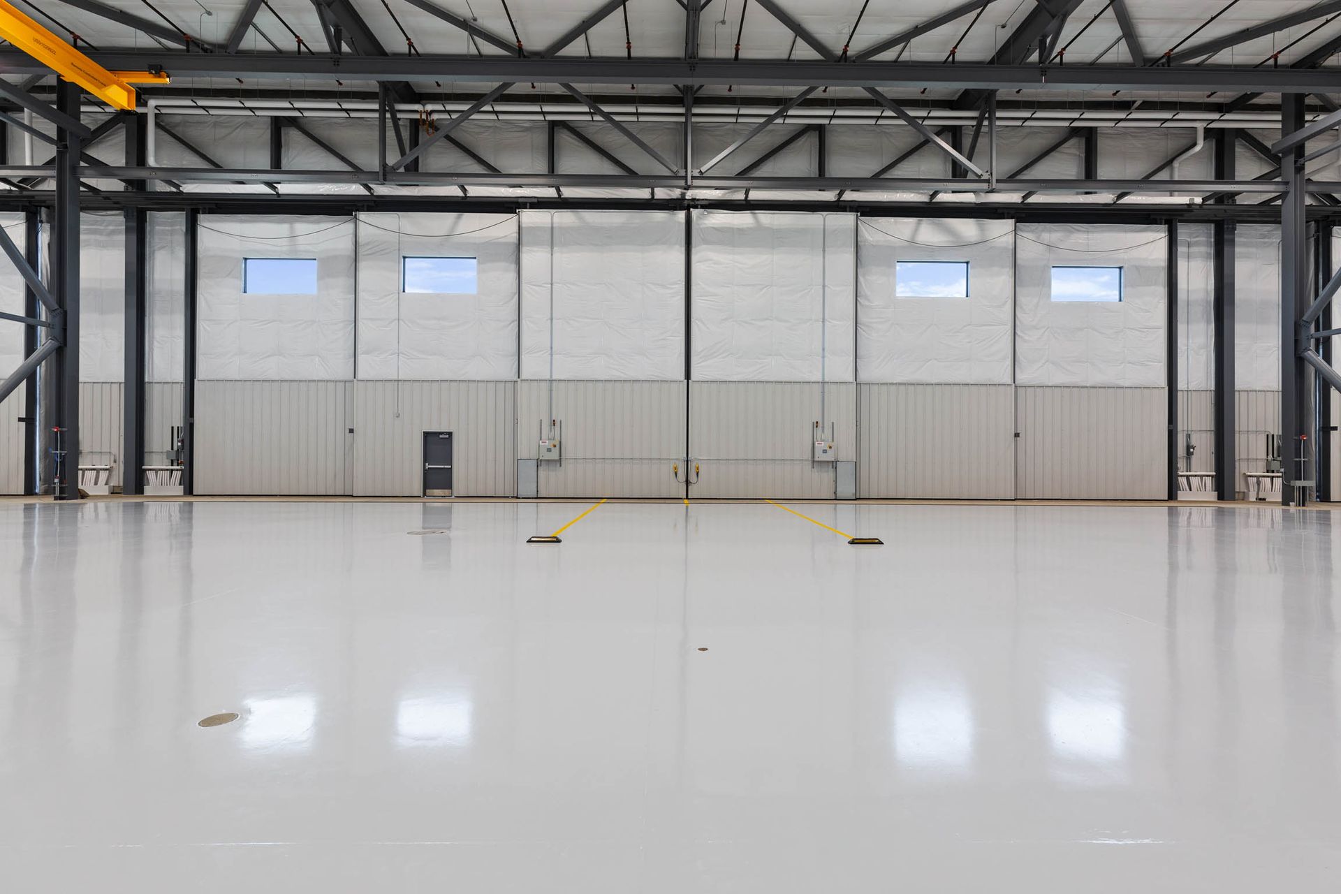 Empty industrial warehouse with a shiny, light gray floor and white walls. Black support beams are visible.