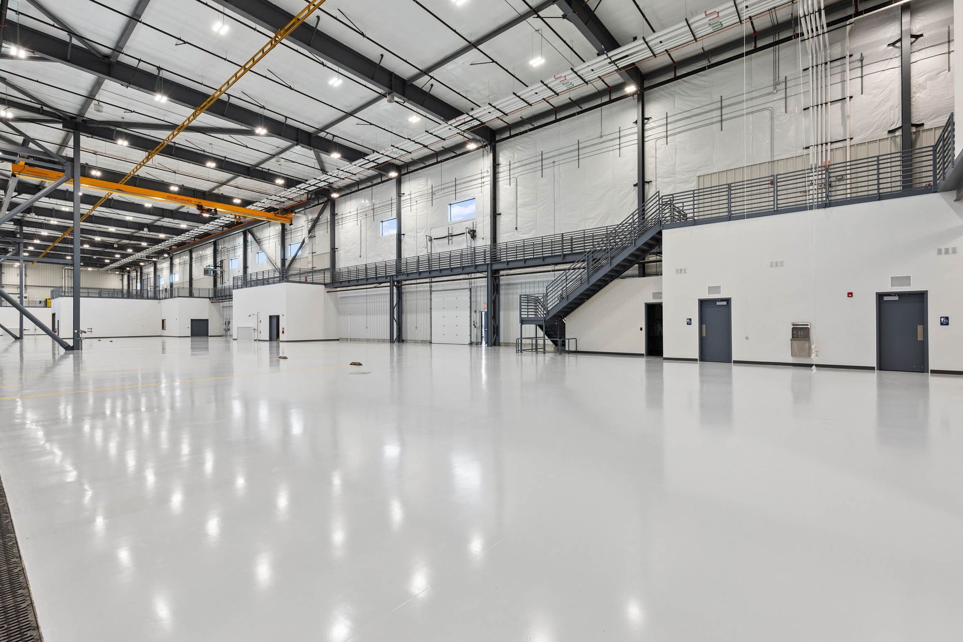 Large, empty industrial building with a shiny white floor. Overhead crane, stairs, and doors visible.