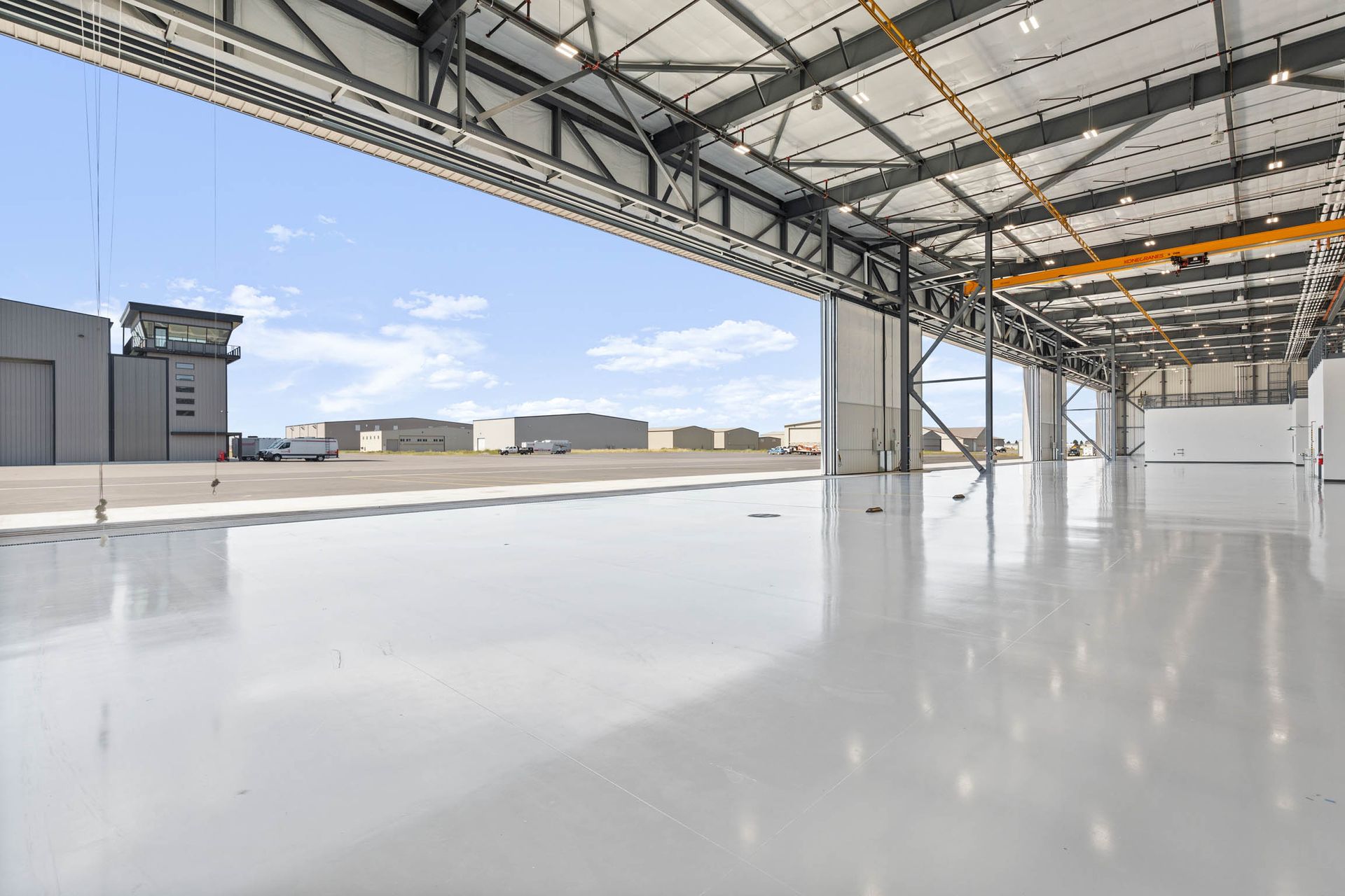 Inside of an aircraft hangar with a reflective, grey floor, open to the tarmac and control tower.