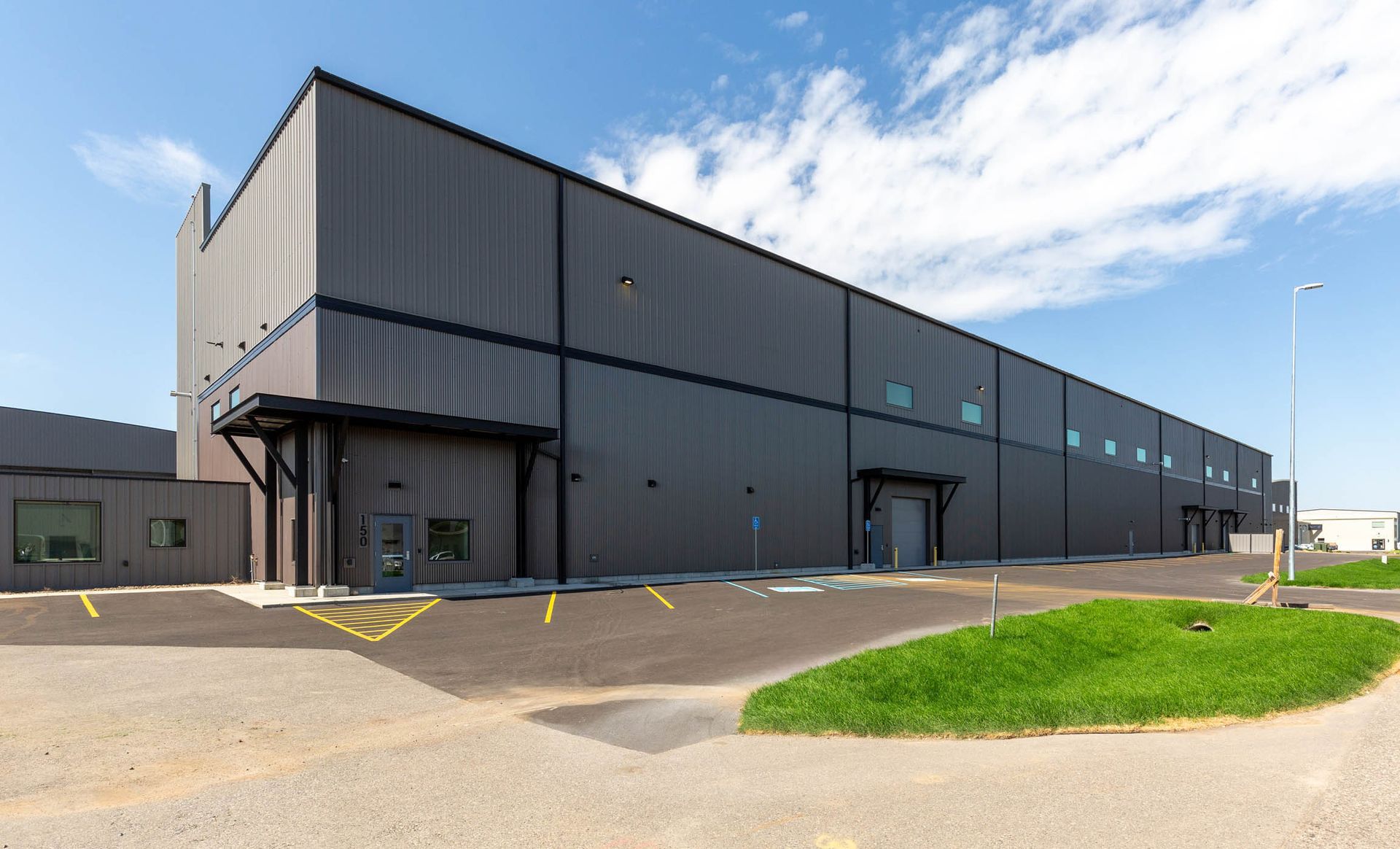 Dark gray industrial building with loading docks, grass, and clear blue sky.