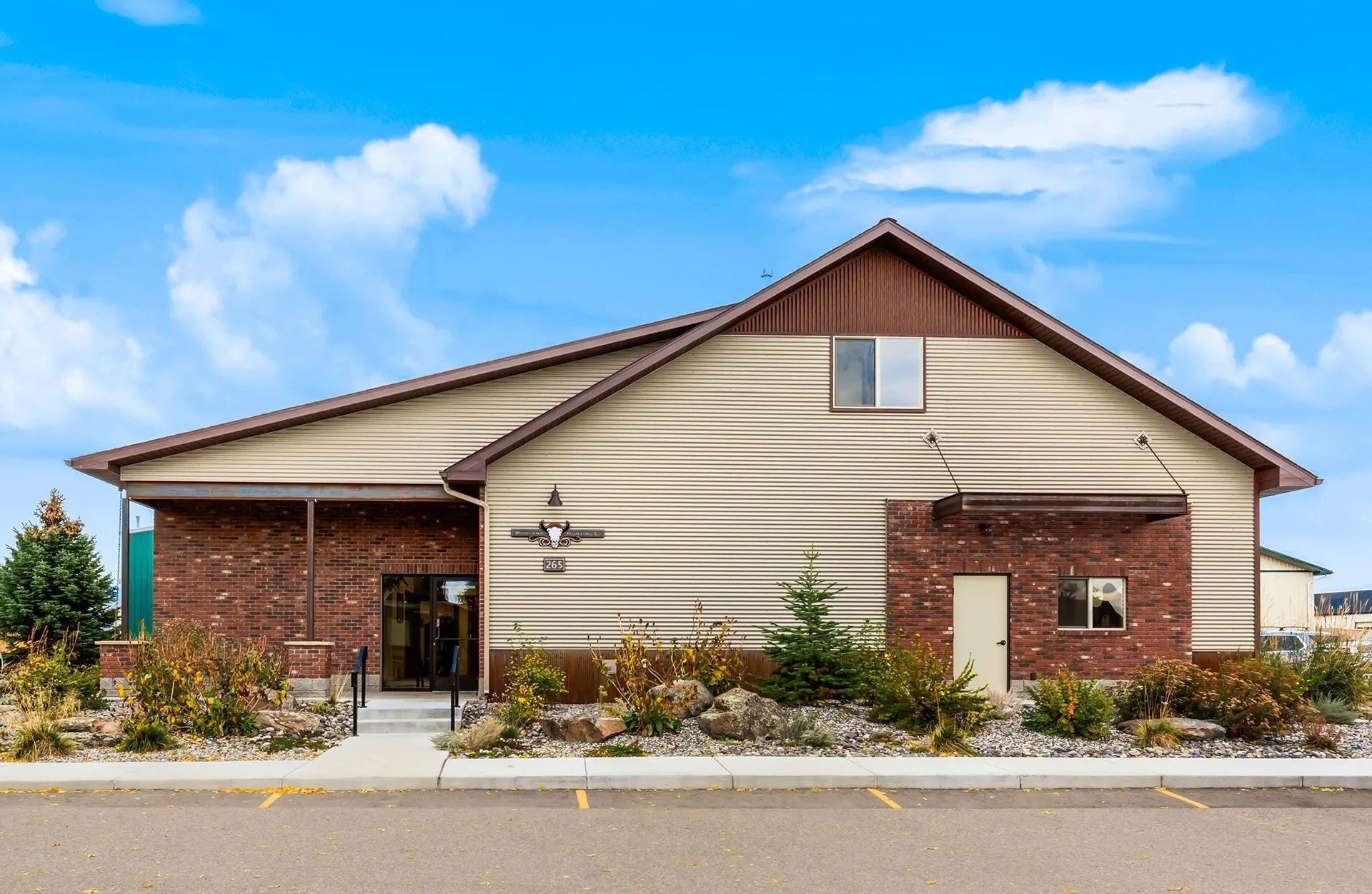 A one-story building with tan siding, brick accents, and a brown gabled roof under a blue sky with clouds.