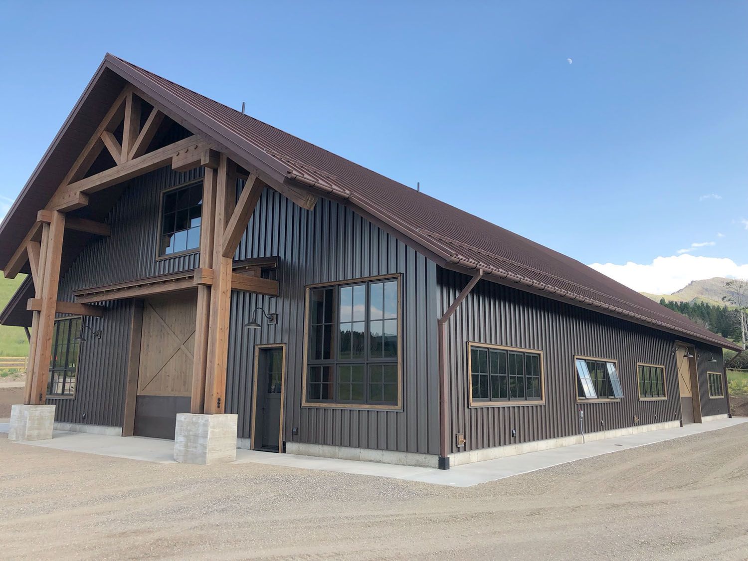 Modern barn-style building with dark metal siding, large windows, and heavy timber framing under a clear blue sky
