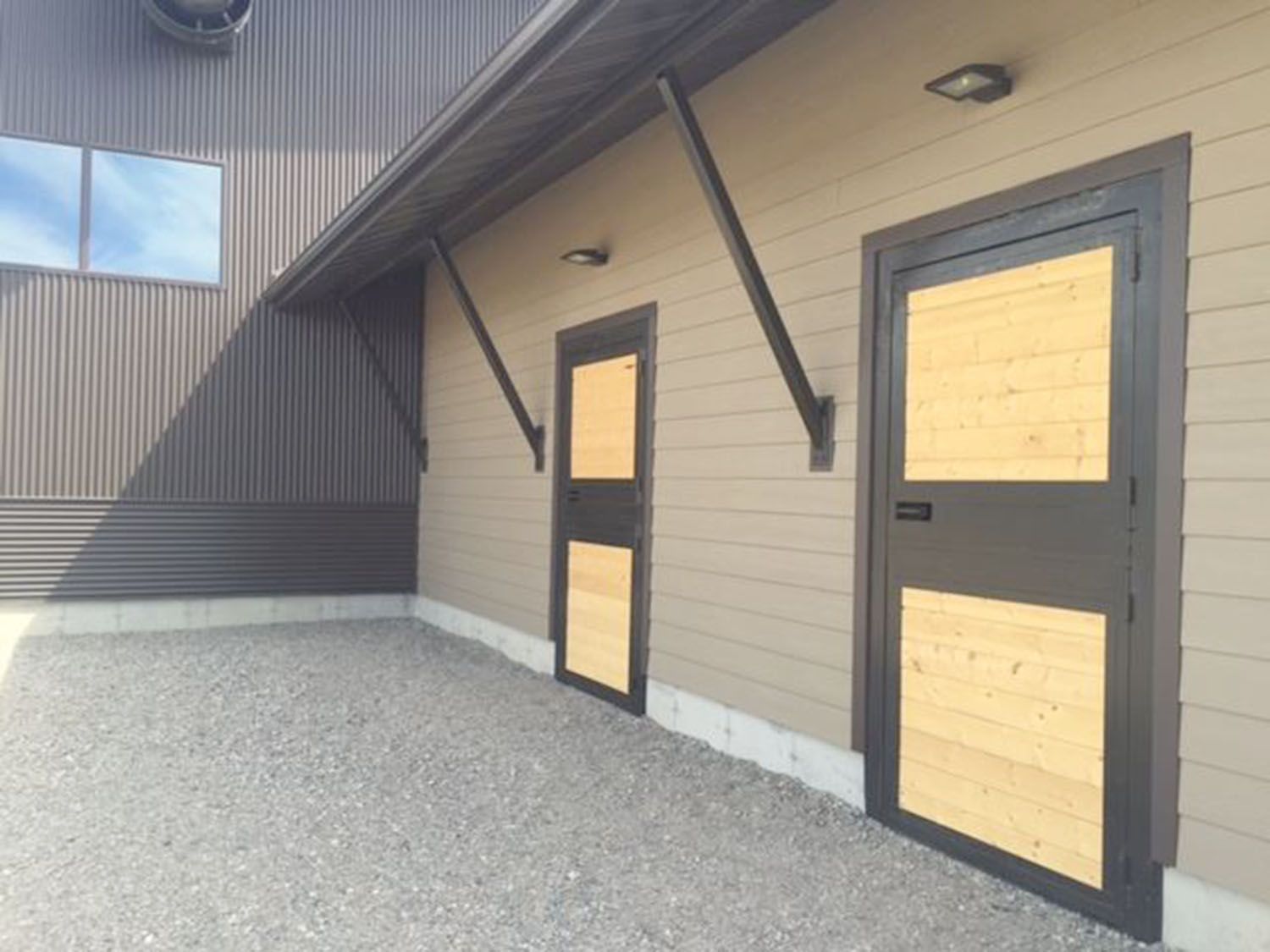 Tan-colored building exterior with two brown, paneled doors and dark metal diagonal braces on a gravel lot