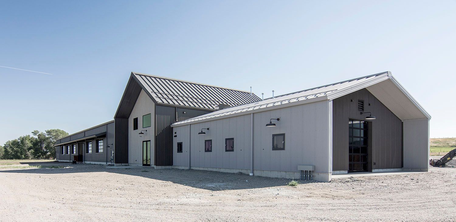 A modern, gray metal industrial or farmhouse-style building with a pitched roof, situated on a gravel lot under blue sky
