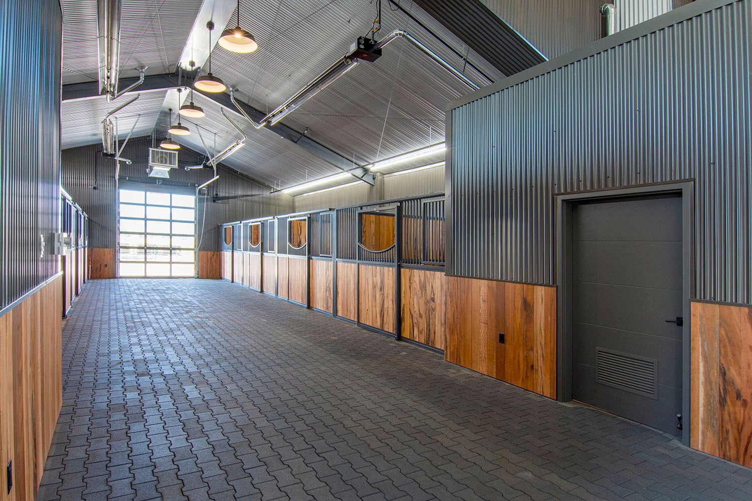 Modern horse stable aisle with wooden stalls, corrugated metal walls, grey brick flooring, and arched ceiling lights