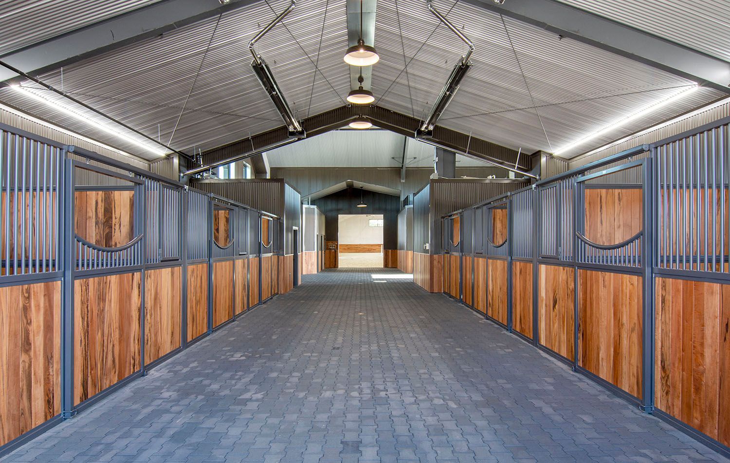 A wide, indoor horse stable aisle with wooden stalls, metal gates, and a paved floor under a high, lit vaulted ceiling