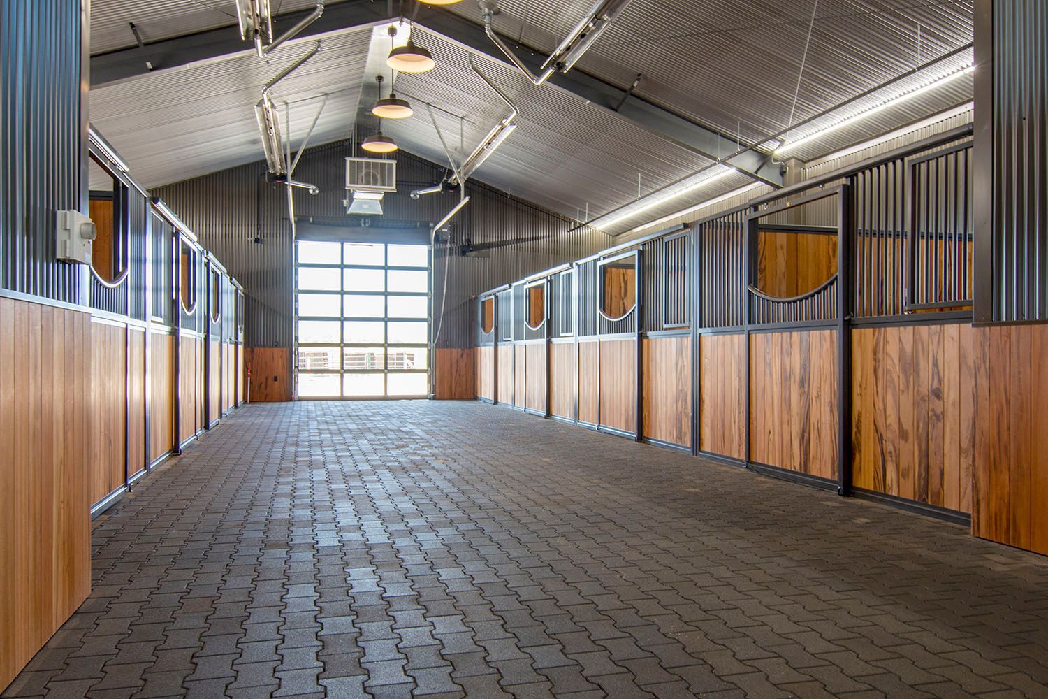 Empty stable aisle with rows of wooden horse stalls, corrugated metal walls, and paved stone flooring under bright lights