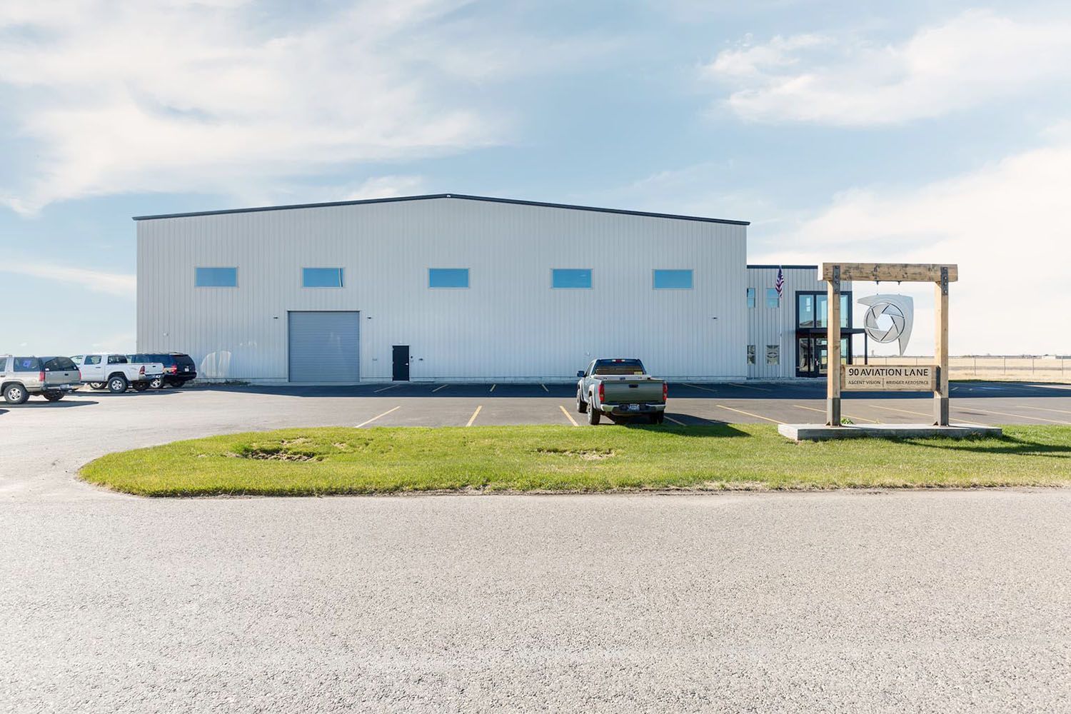 A modern, white industrial warehouse building with a parking lot, a gray truck, and a wooden sign in front