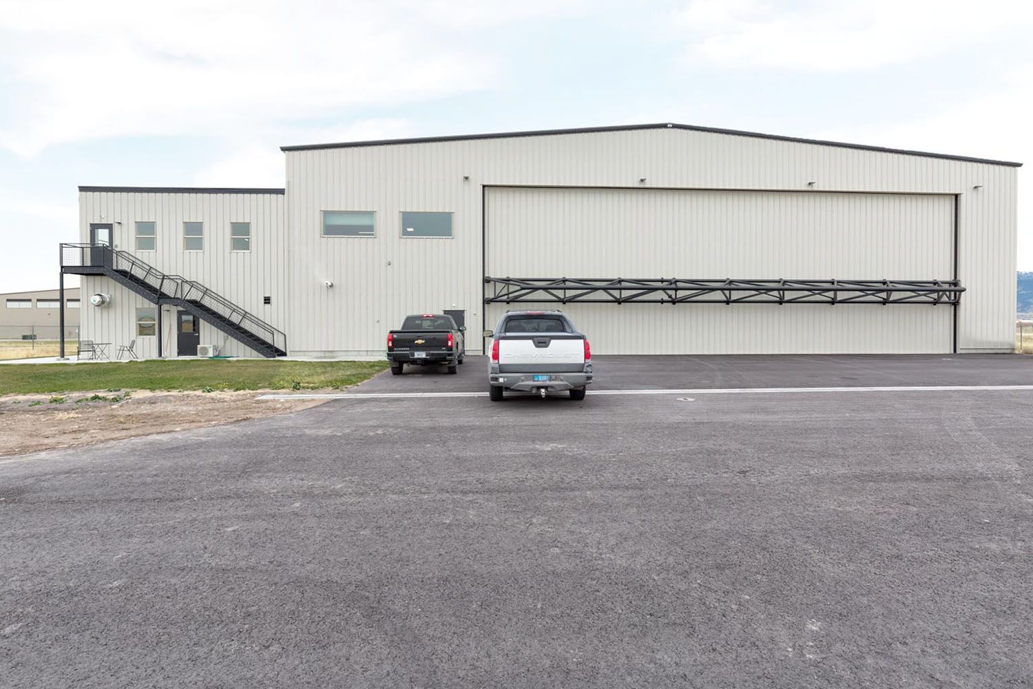 A light-colored metal hangar building with exterior stairs and two pickup trucks parked in front on an asphalt lot