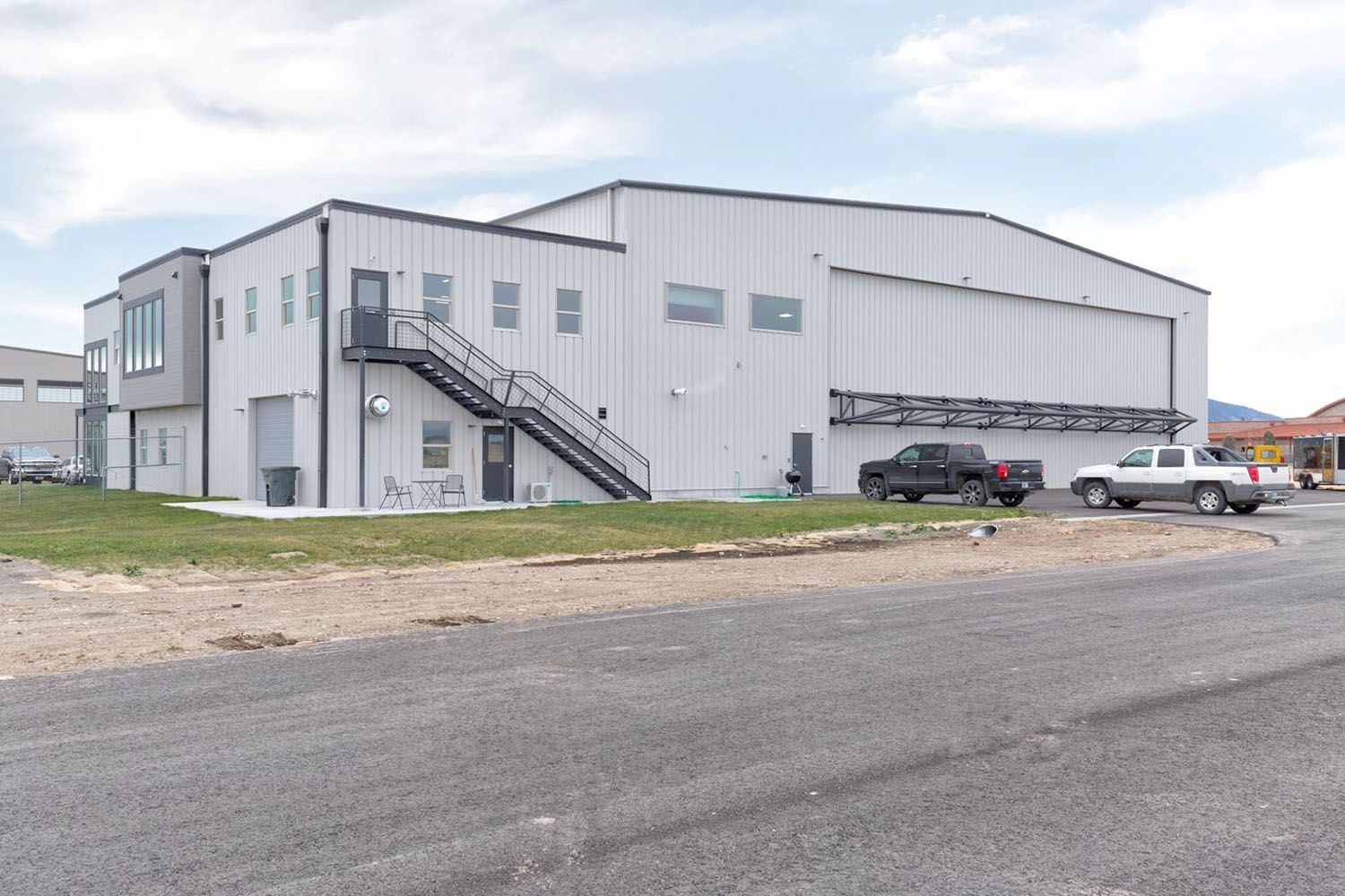 A white hangar building with an exterior metal staircase and two pickup trucks parked in front on a gravel lot